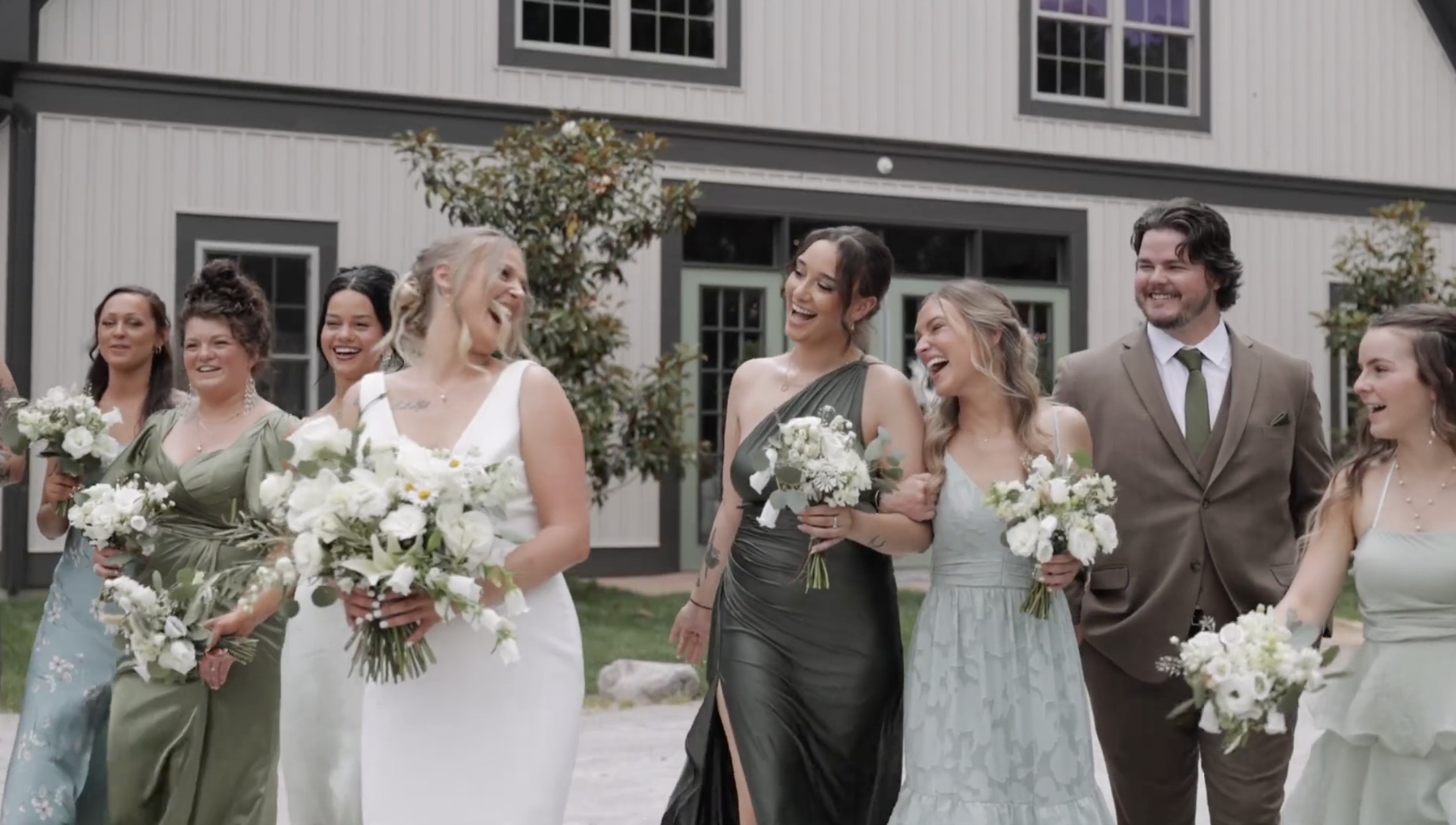 A group of women and one man in wedding attire, smiling and walking outdoors with bouquets of white flowers in front of a modern house with large windows and trees.