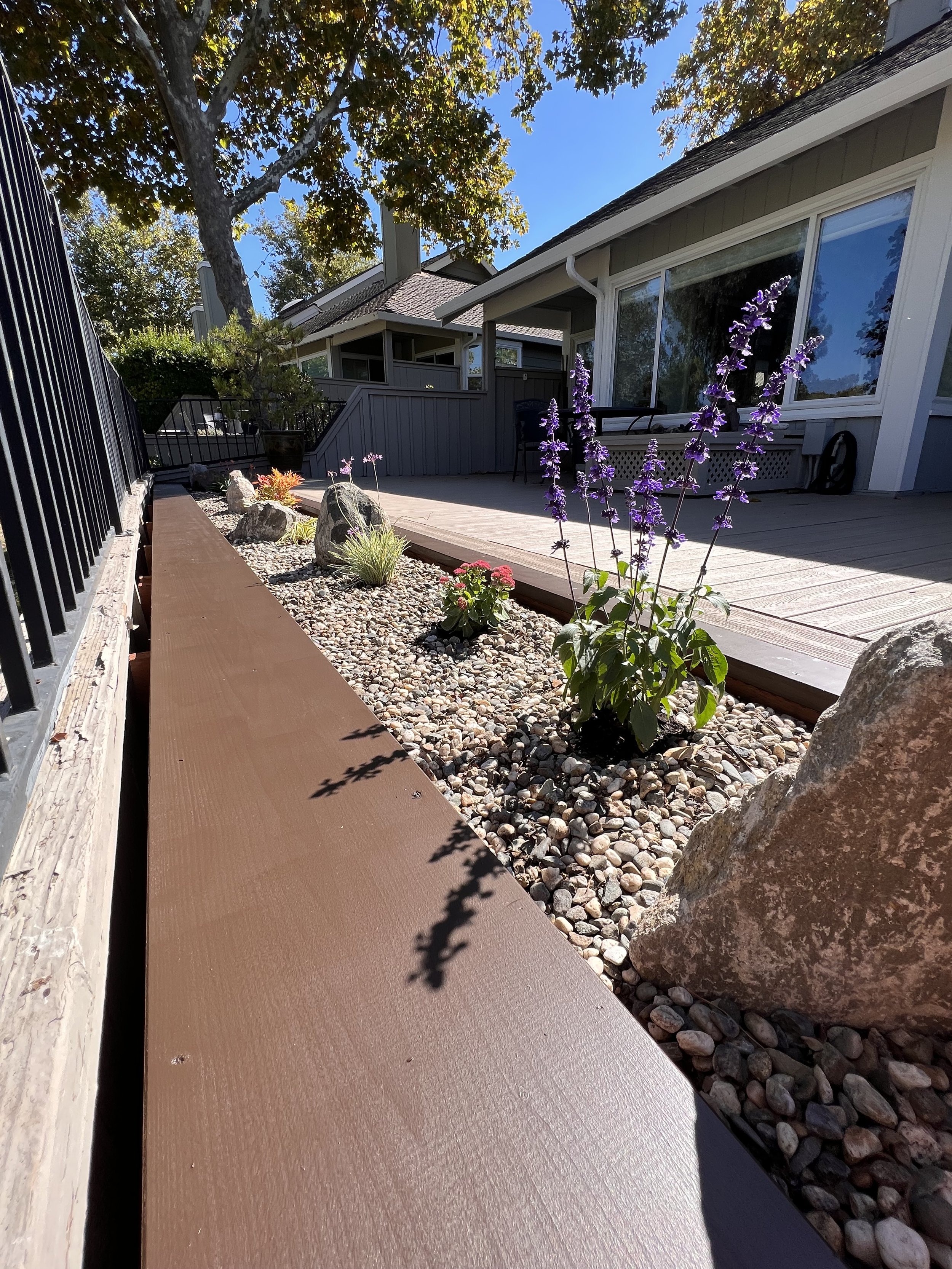 A backyard patio with a wooden deck, adjacent to a flower bed with purple and pink flowers, rocks, and large stones, next to a house with large windows. There is a tree providing shade and a bright blue sky.