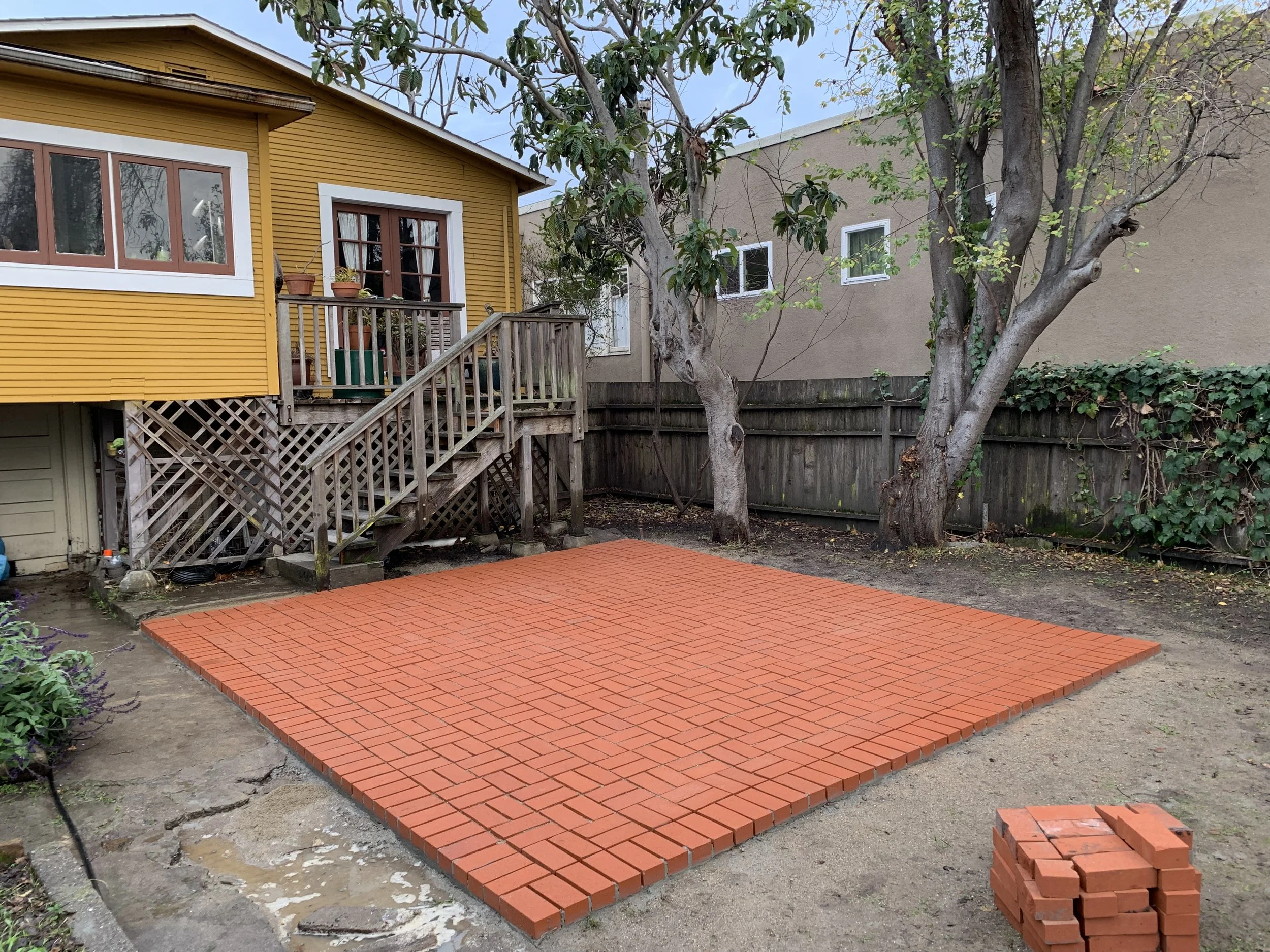Backyard with a newly built red brick patio, a yellow house with a staircase, and trees along a wooden fence.