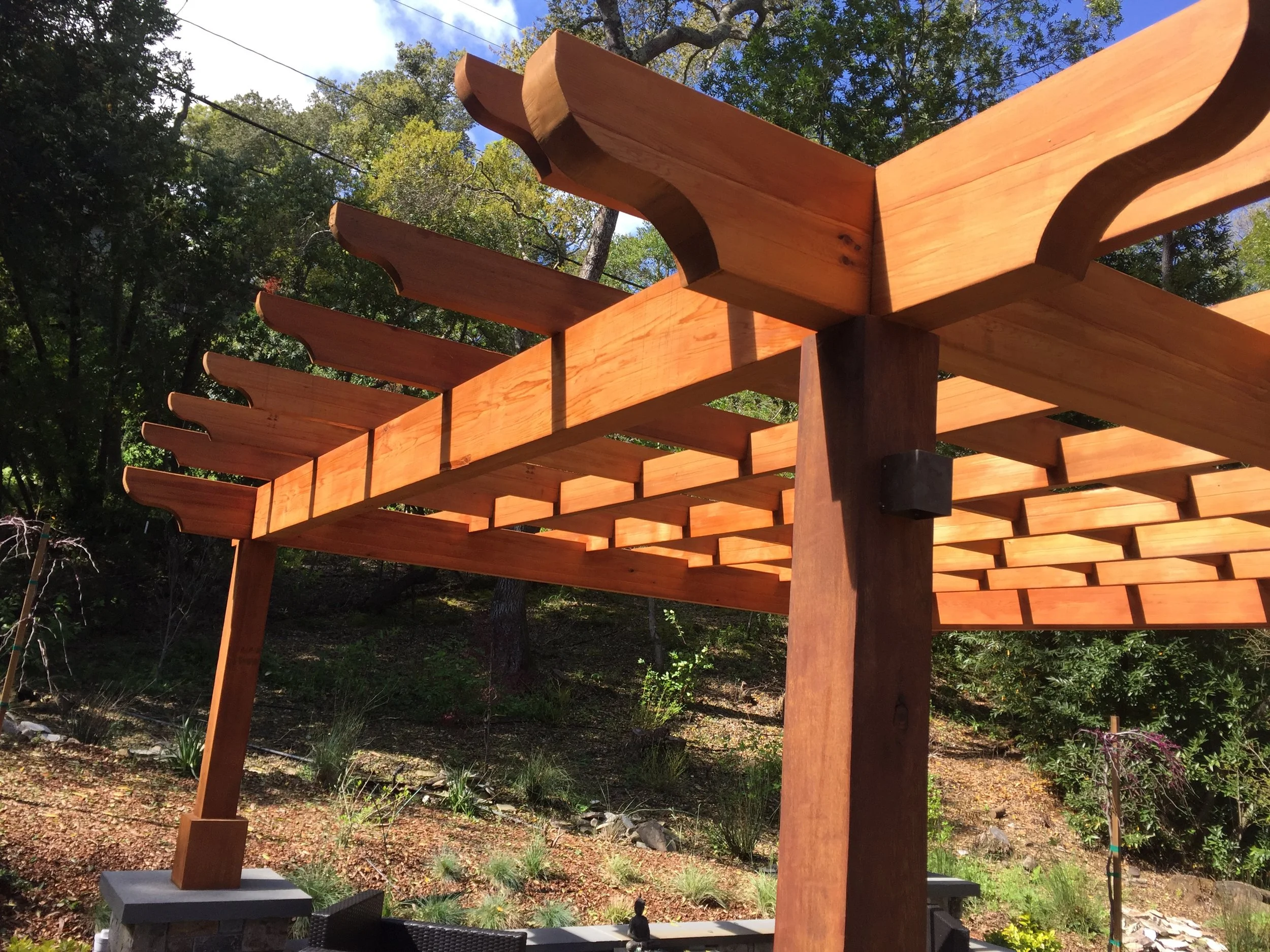 Wooden pergola structure outdoors with beams and lattice roof, surrounded by trees and landscaping.