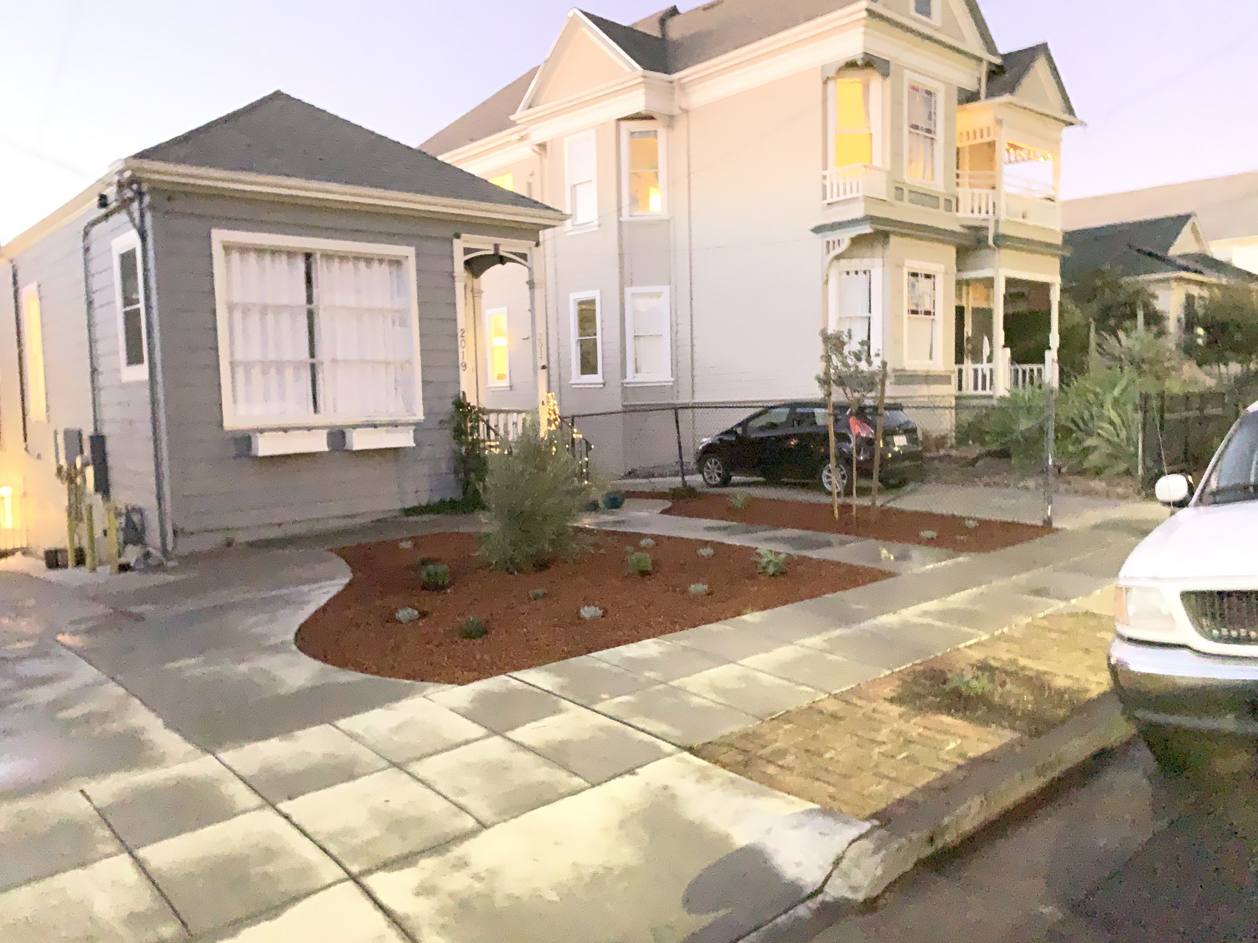 Residential neighborhood with a sidewalk, a small garden area with plants, and two houses, one with a bay window and the other with a covered porch and multiple stories, during evening time.