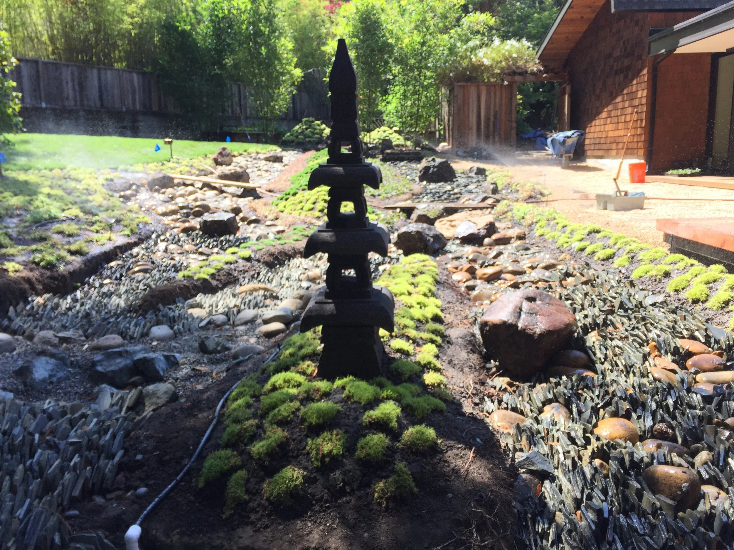 Landscaping in progress, with decorative rocks, moss-covered plants, a pond, and a partially constructed garden bed near a house with a brick exterior.