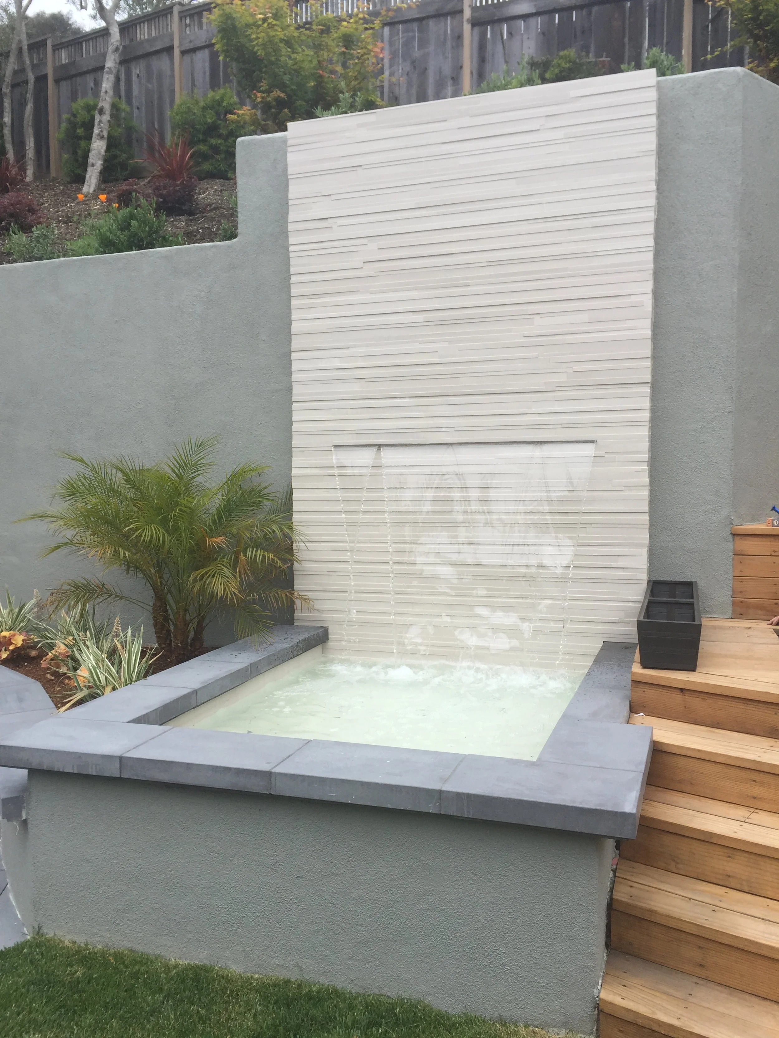 Outdoor hot tub with water feature, white decorative wall, wooden stairs, and landscaped garden with plants and trees.