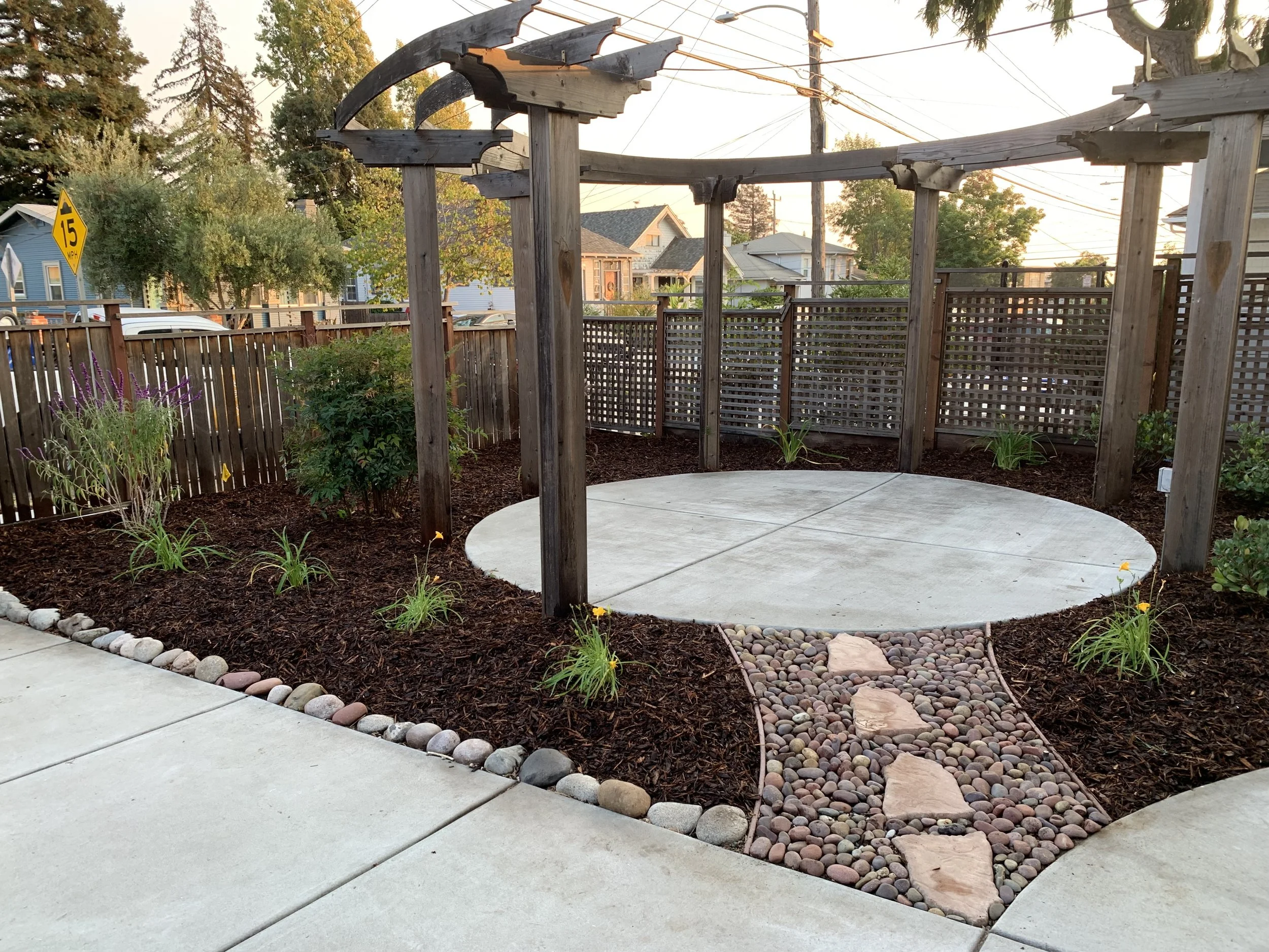 A backyard garden area with a circular concrete patio, a pebble path with large stepping stones leading to it, landscaped flower beds with small plants and mulch, a wooden pergola frame, and a wooden fence in the background. The yard is in a suburban