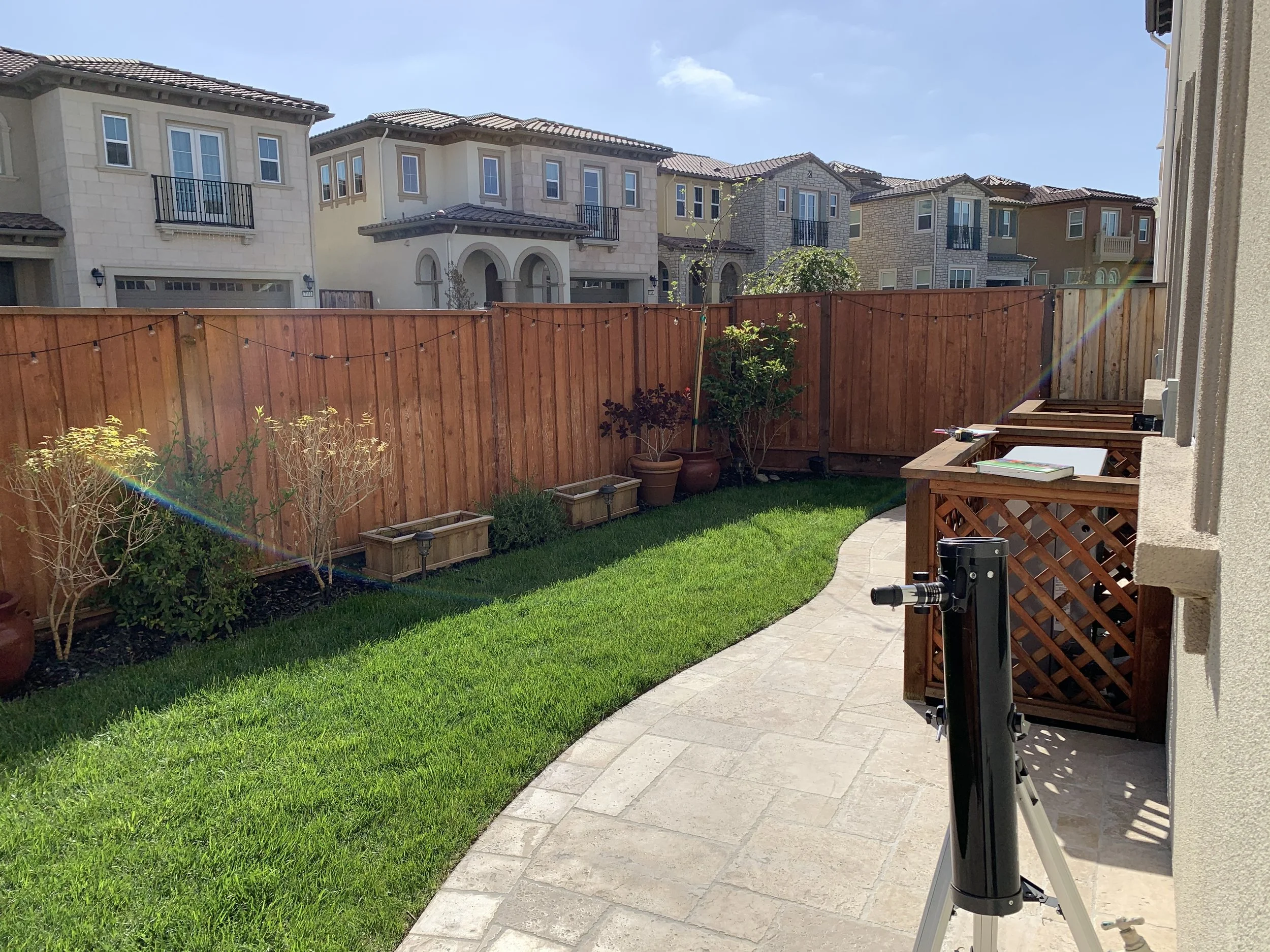 Backyard with a curved brick pathway, green lawn, and potted plants along a wooden fence. There is a telescope on a tripod near the house on the right. In the background, multi-story houses with tile roofs are visible against a partly cloudy sky.