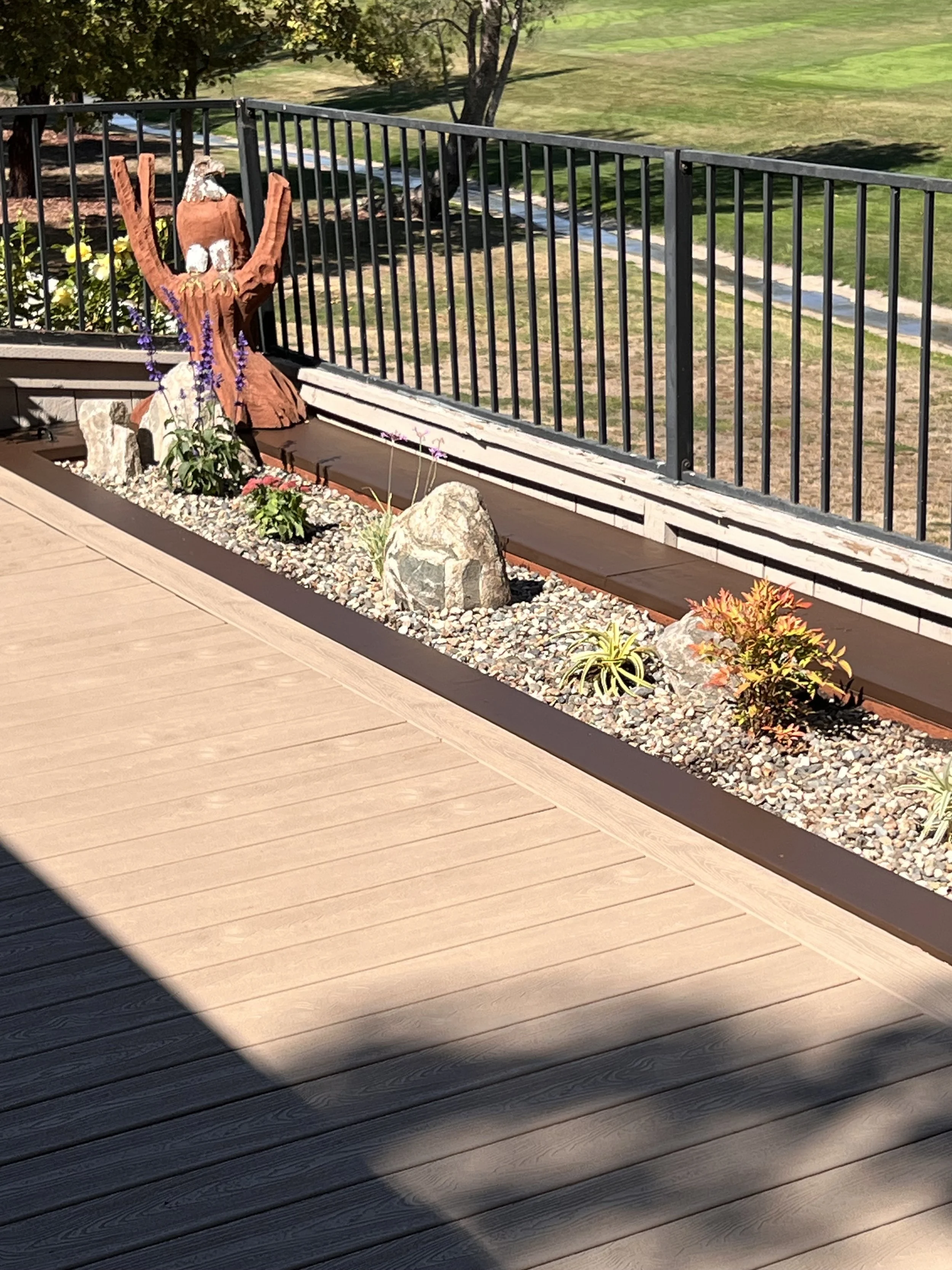 Decorative garden bed with a wooden eagle sculpture, rocks, and various plants, bordered by a yard with grass and trees, and a black metal railing on a wooden deck.
