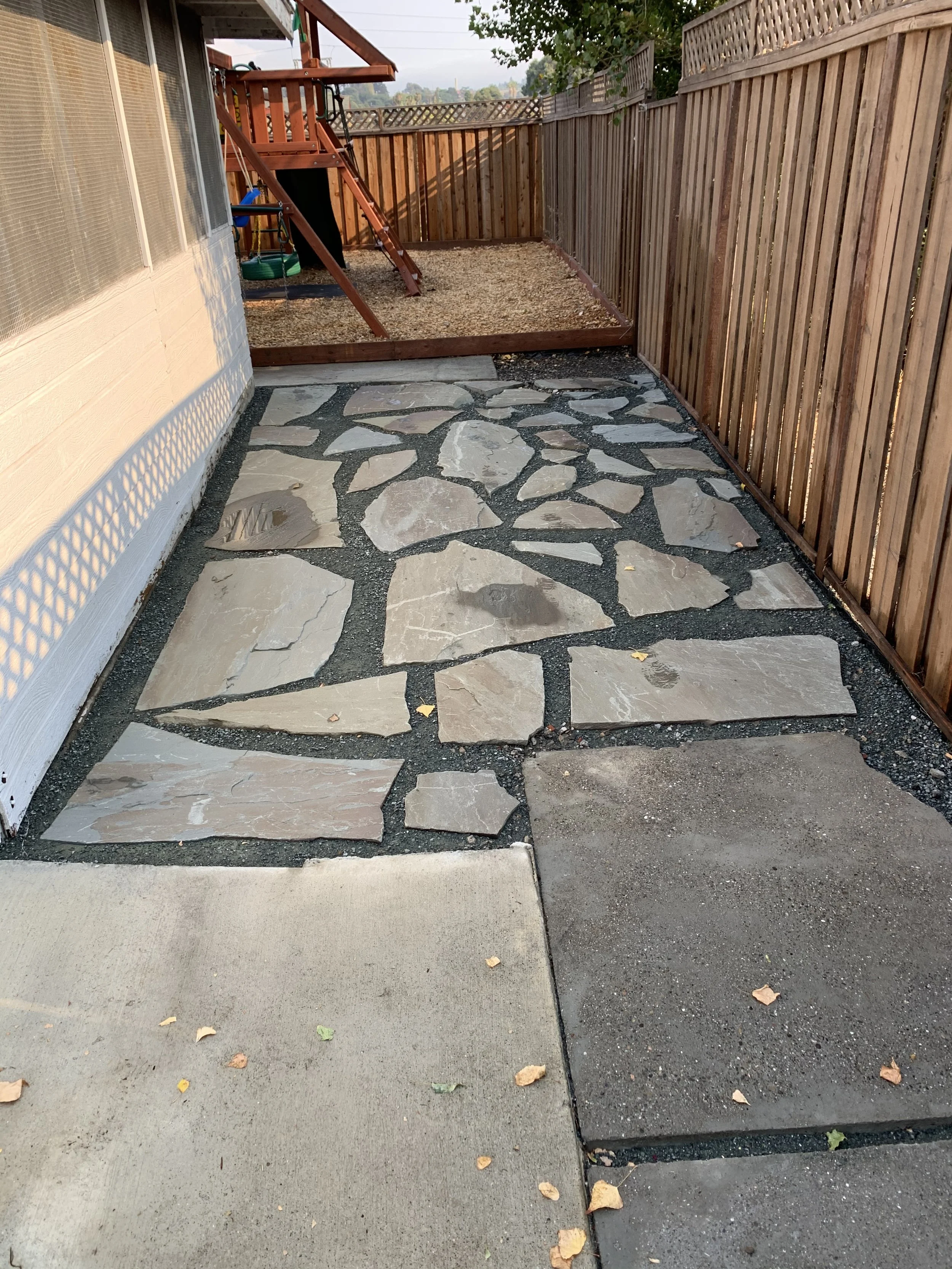 A backyard patio with a concrete slab, a stone pathway, a wooden fence, and a small fenced-in area with a wood deck and a swing set.