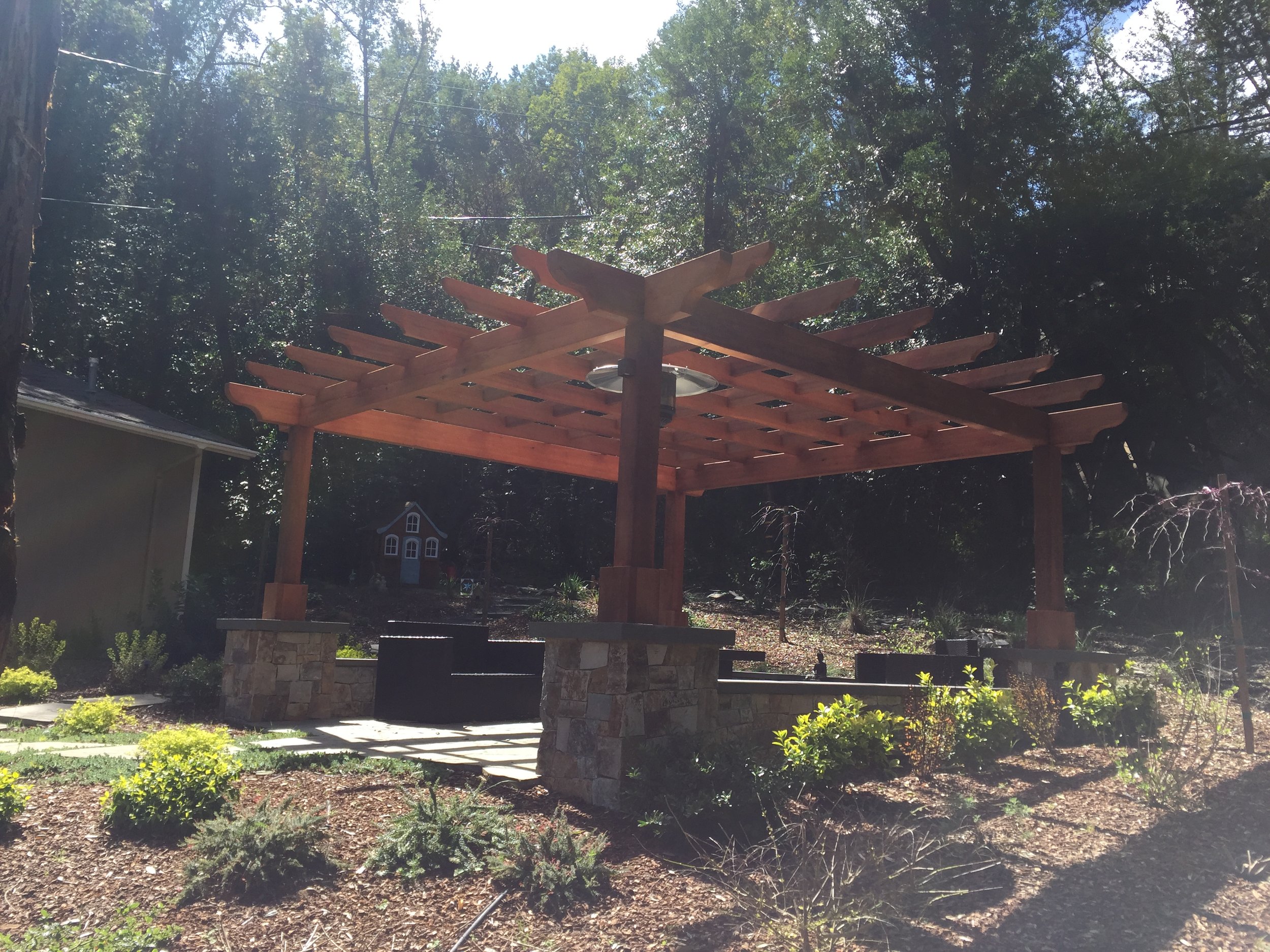 Wooden pergola with stone base in a garden, surrounded by bushes and trees, with a small playhouse in the background.