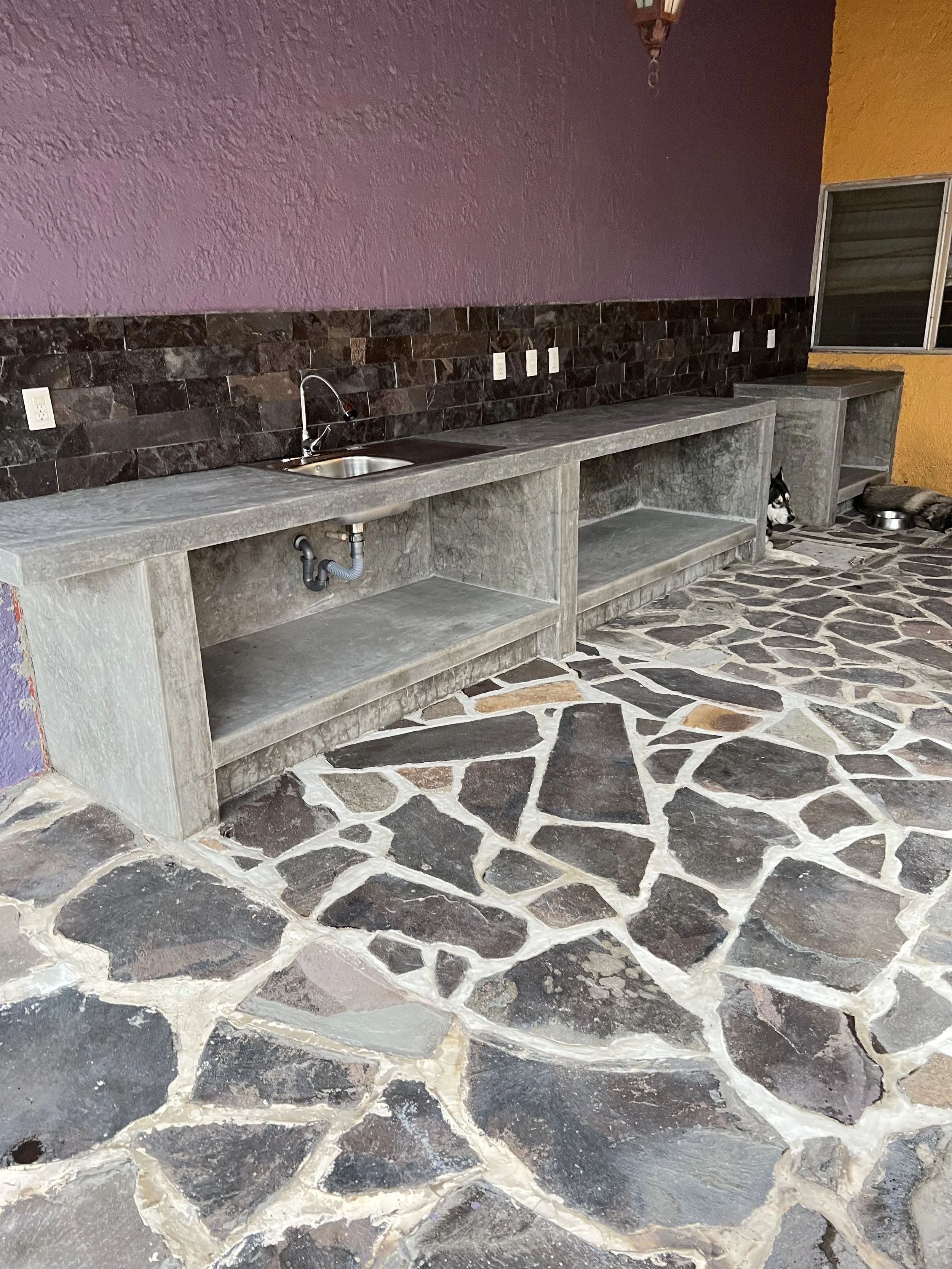 Concrete outdoor kitchen with a single sink, open shelving, and a stone floor. There are two dogs resting near the wall, and a window to the right.