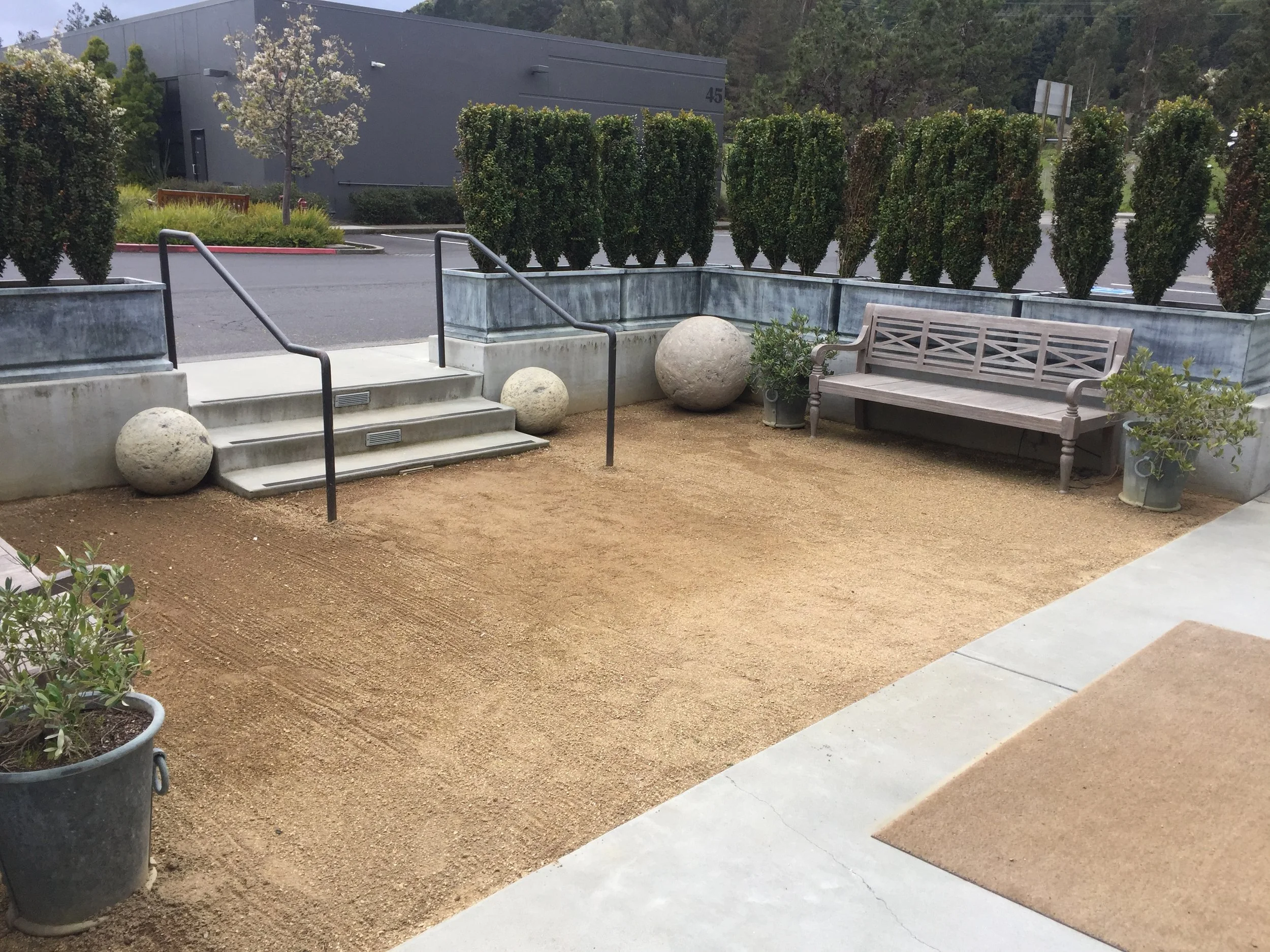Empty outdoor patio area with a small runway of sand, a wooden bench, potted plants, decorative large spherical stones, and a staircase with black handrails. There are shrubs and trees in the background.
