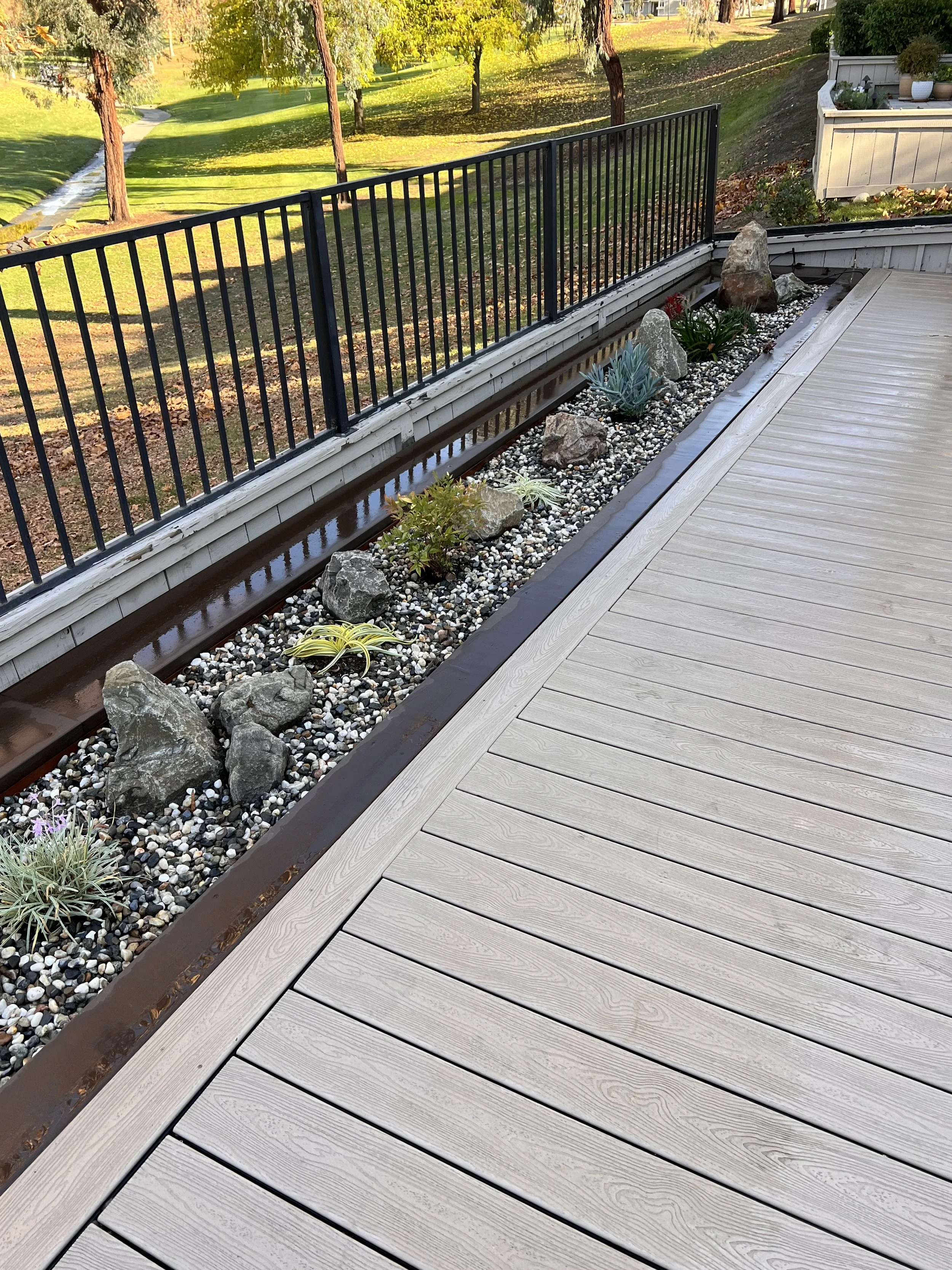 A garden with a wooden deck and a narrow plant bed separated by a metal railing, containing rocks, succulents, and small plants, overlooking a grassy park with trees.