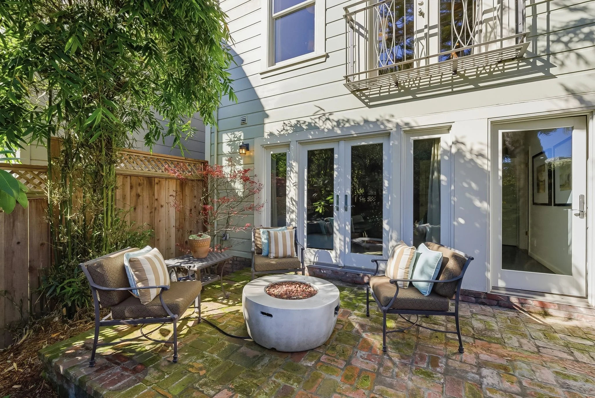Patio area with four chairs, a circular fire pit, and potted plants, surrounded by a brick floor and beige house exterior with sliding glass doors and a small balcony.