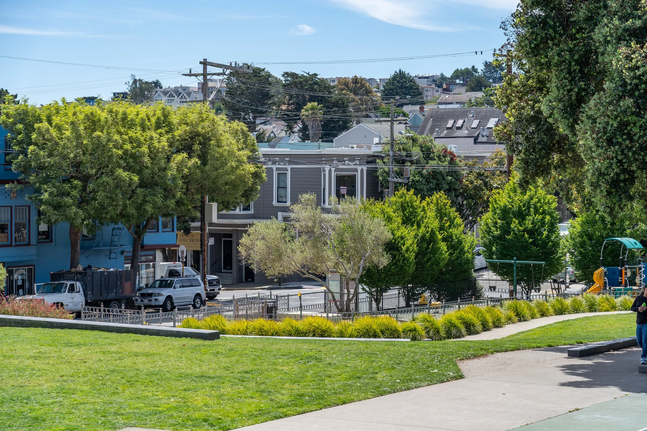 A park with a walkway, green grass, and bushes, with residential buildings and trees in the background under a clear sky.