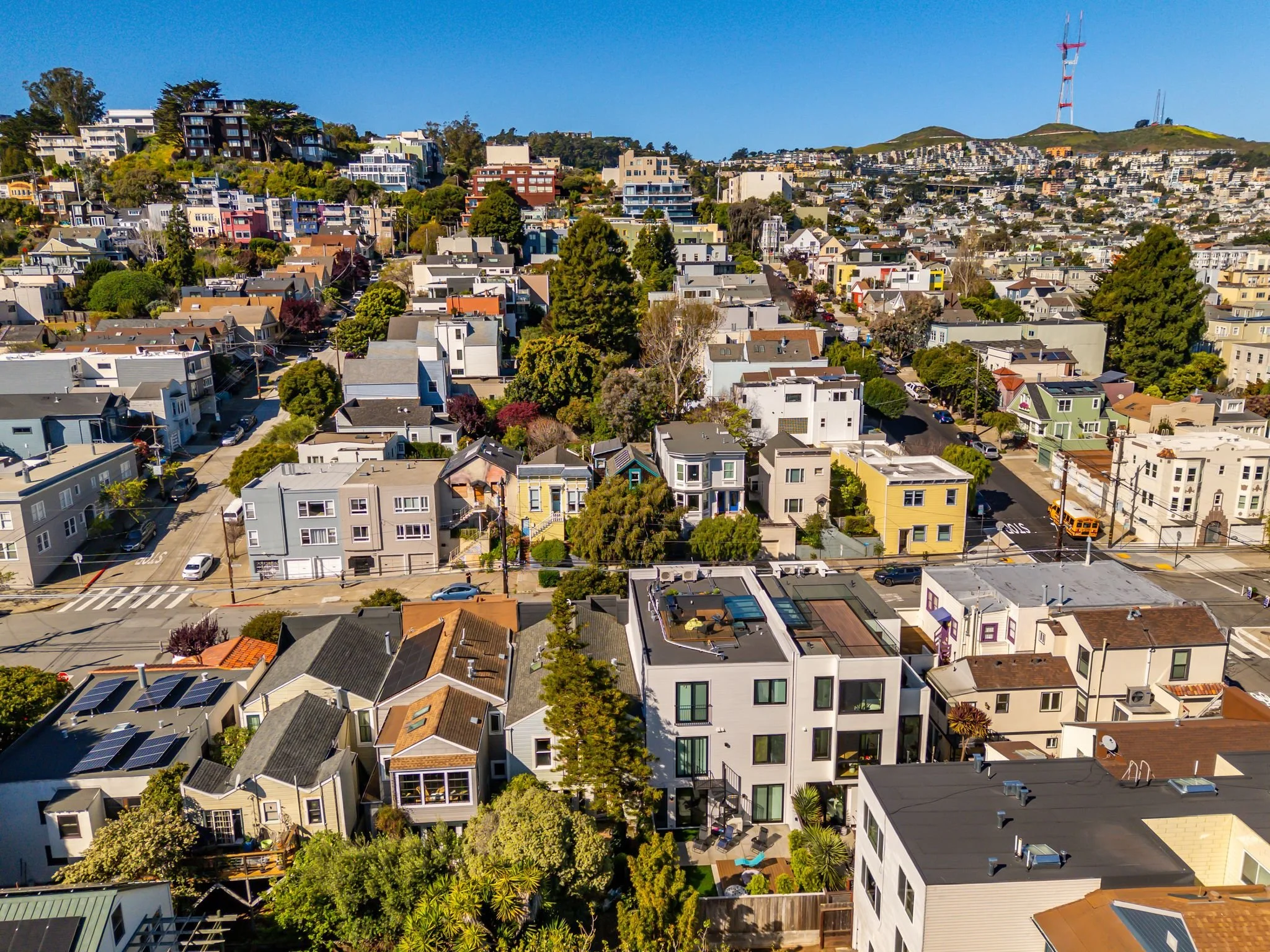 Aerial view of a hillside neighborhood with colorful houses, trees, and a radio tower on top of the hill in the background under a clear blue sky.