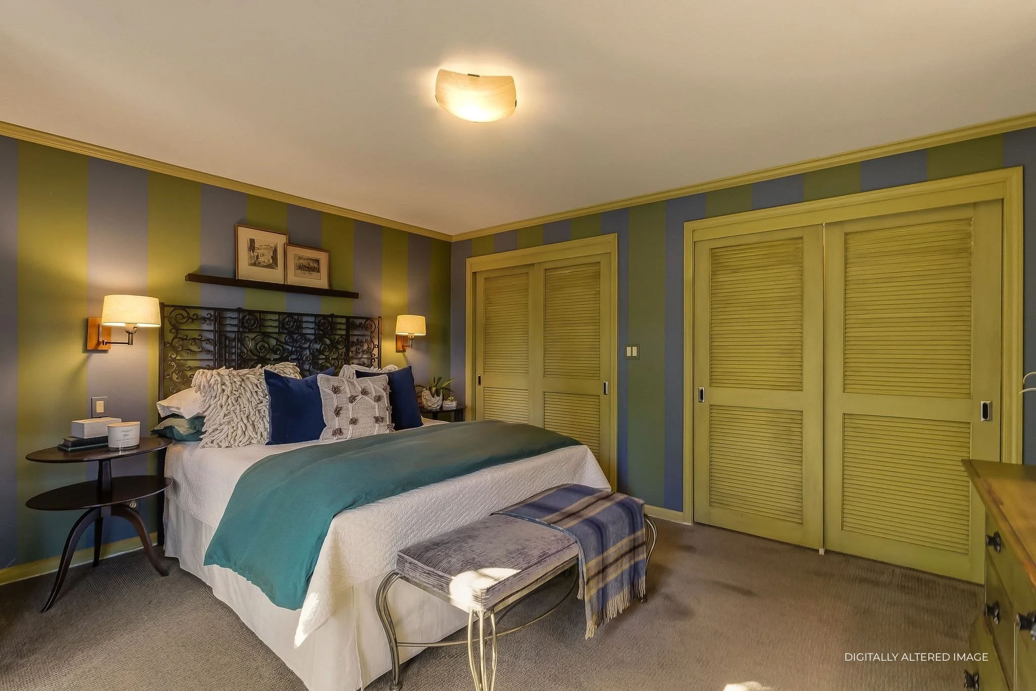 A bedroom with green and blue striped walls, a bed with many pillows, two wall-mounted lamps, a bench at the foot of the bed, and a closet with louvered doors.