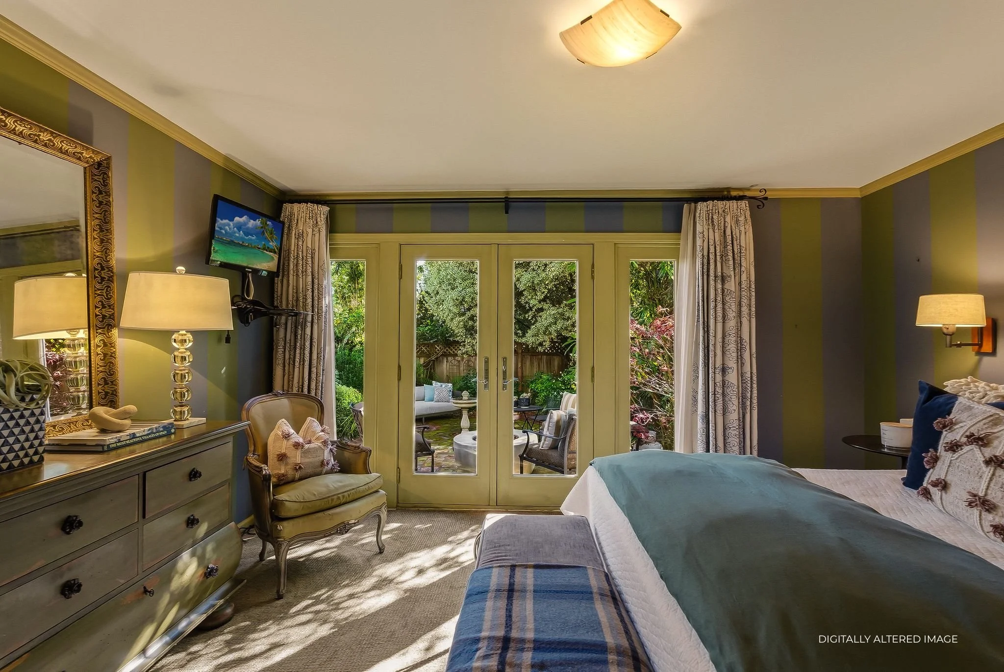 A bedroom with striped green and gray walls, a large bed with pillows and a blue blanket, a pairing of French doors with curtains leading to an outdoor patio, a vintage-style armchair, a dresser with a mirror, lamps, and a small wall-mounted television.