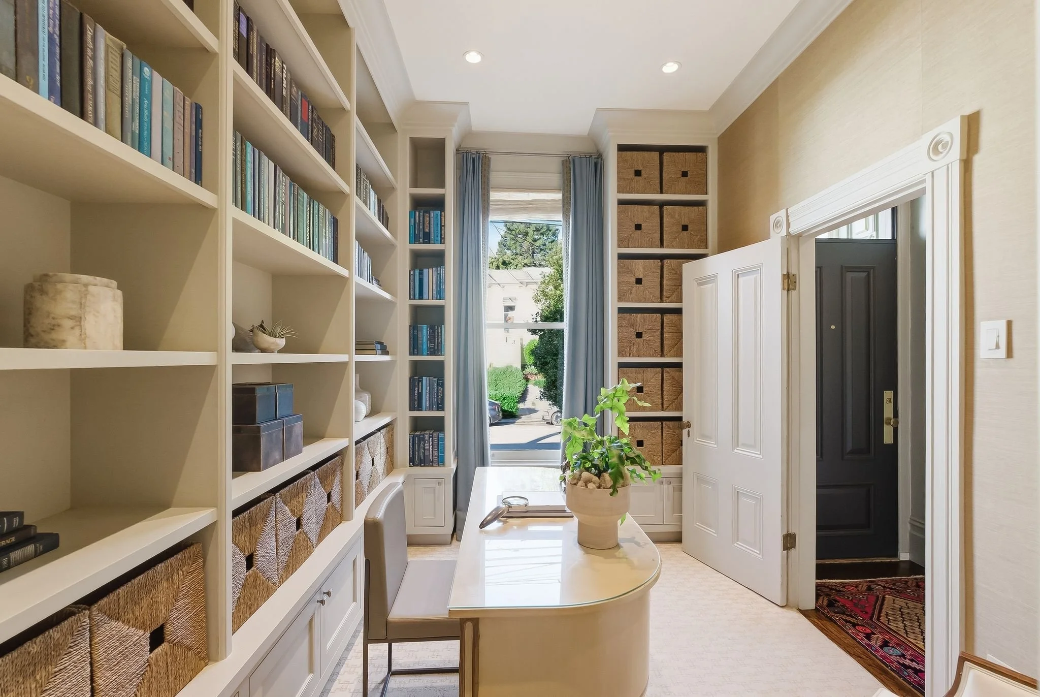 Bright room with built-in white bookshelves filled with books and storage baskets, a white desk with a potted plant, large window with sheer curtains, and a dark front door.