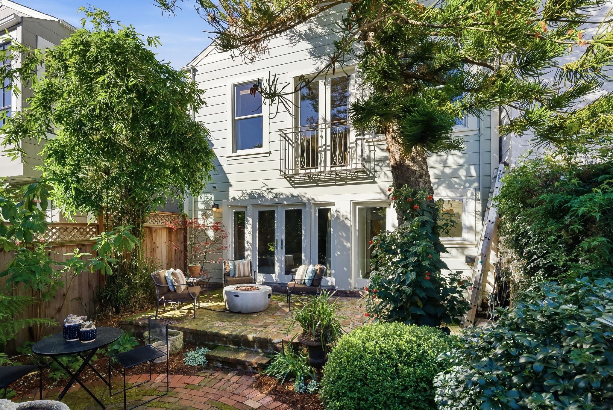 A cozy backyard patio with outdoor furniture, surrounded by lush green trees and plants, attached to a white two-story house with multiple windows and a small balcony.