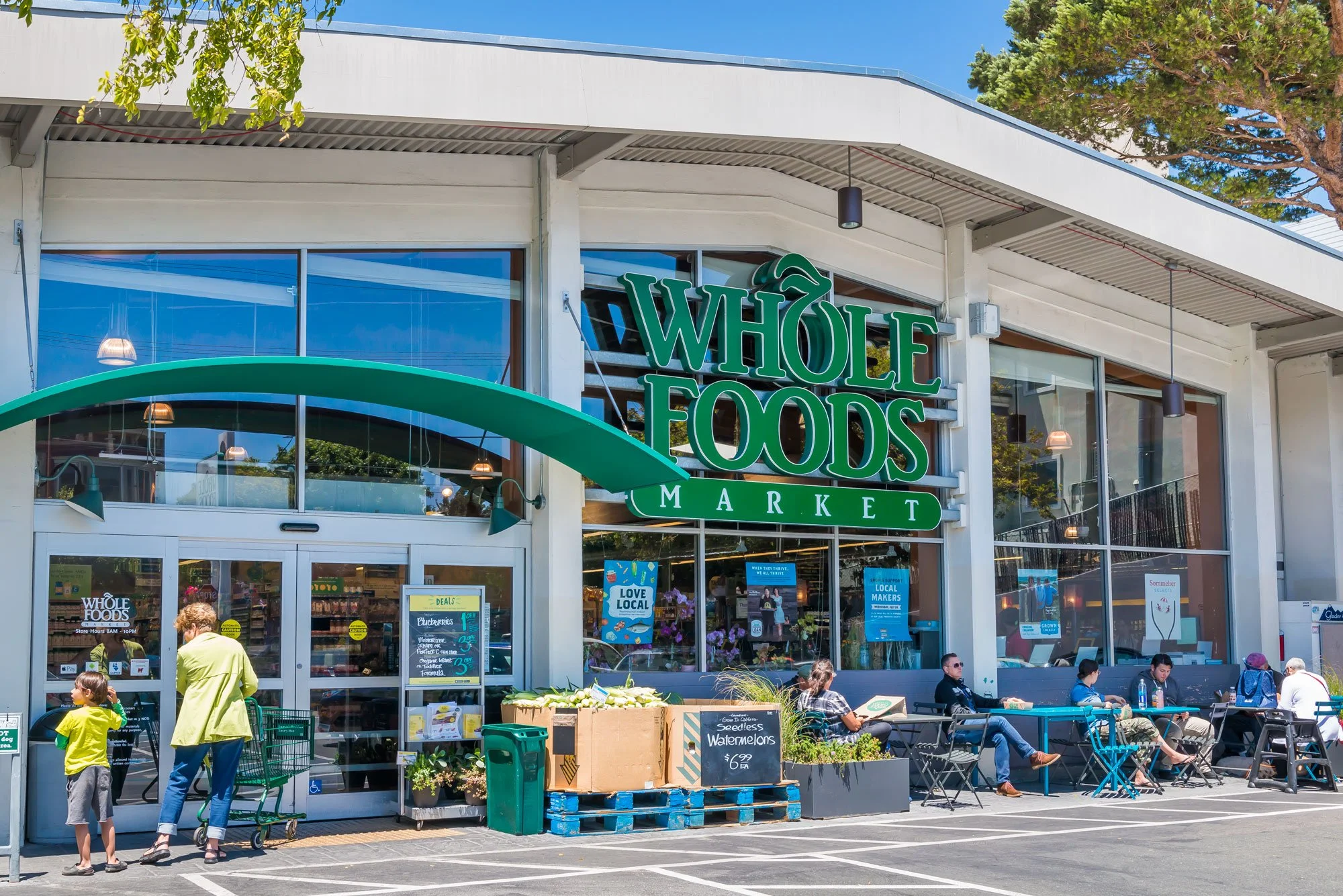 Exterior view of Whole Foods Market with people sitting outside and shopping. Large green sign displays the store name.
