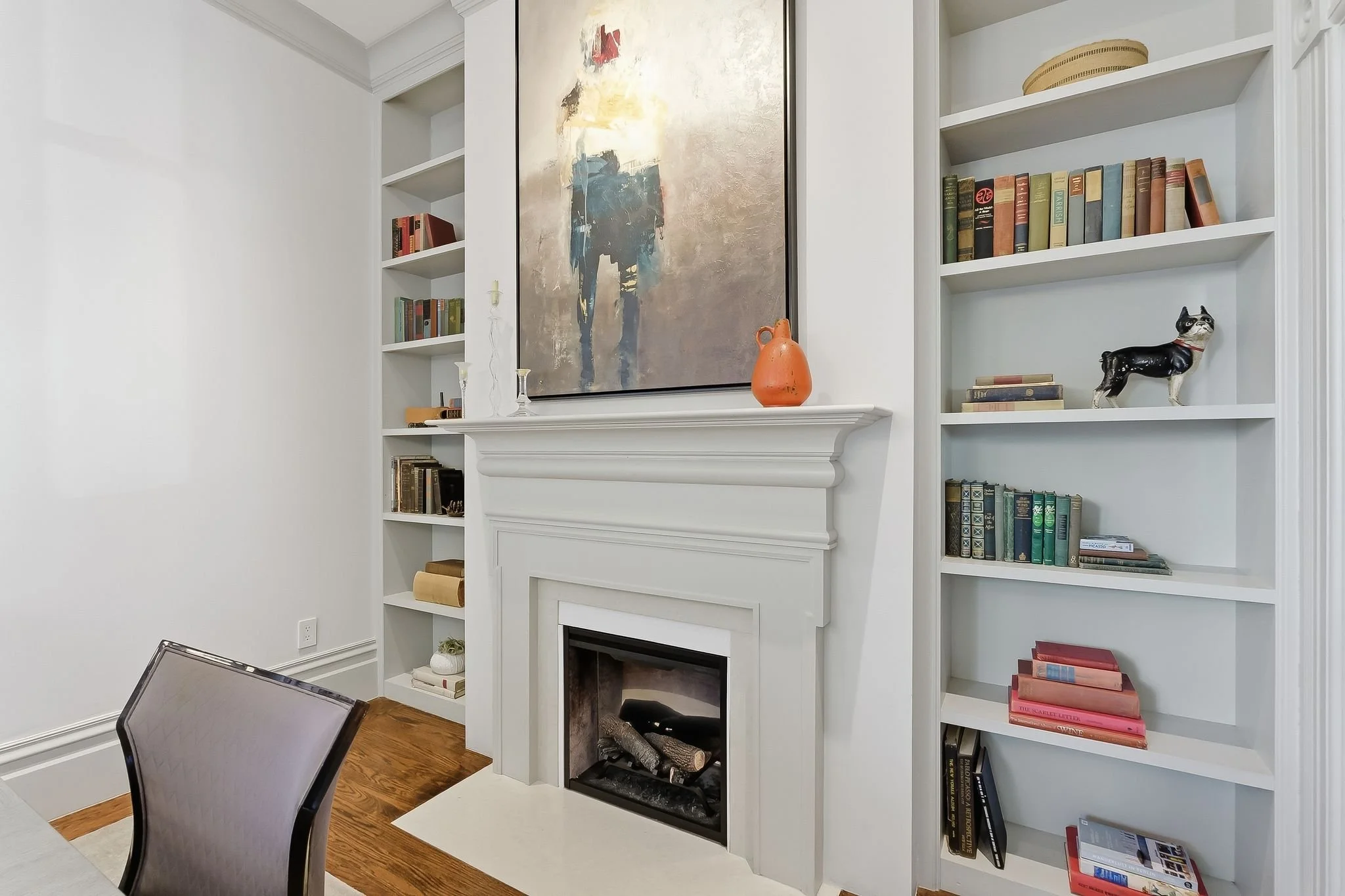White built-in bookshelf with books and decorative items surrounding a white fireplace with a large abstract painting above and an orange vase on the mantel. A chair is partially visible in the foreground.