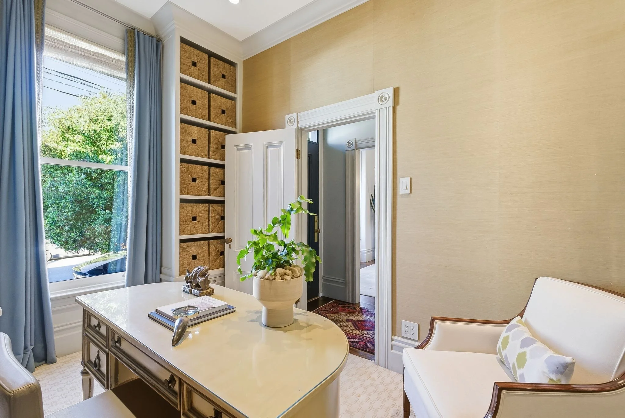 A home office with a window with blue curtains, a white desk with a plant, and a white armchair with a pillow.