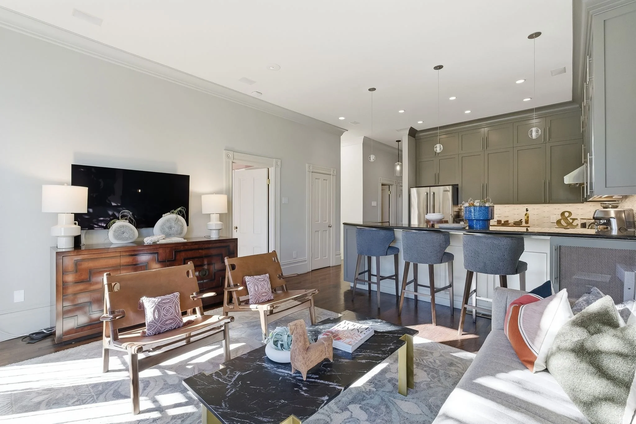 Bright open-concept living room and kitchen with modern furniture, a large flat-screen TV, and a bar area with three barstools.