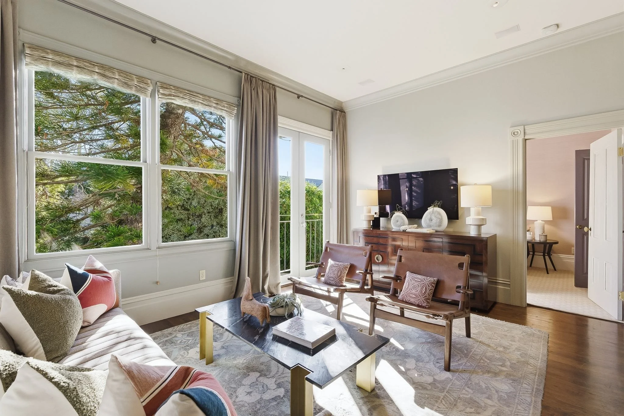 Bright living room with large windows and beige curtains, a wooden sideboard with decorative vases, a flat-screen TV, two table lamps, and a seating area with cushions on sofa and chairs, a coffee table, and a rug.