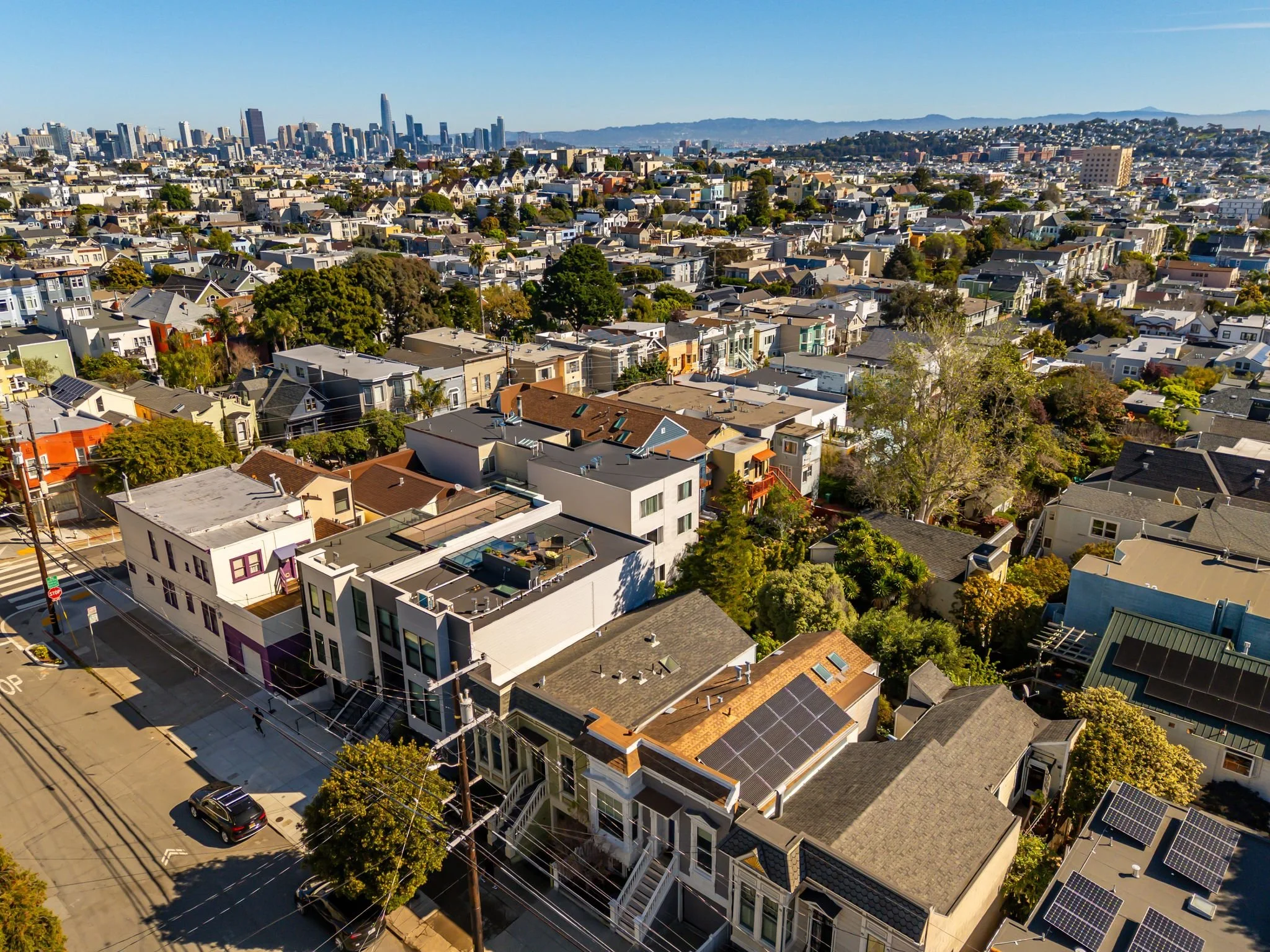 Aerial view of a cityscape with a mix of residential and commercial buildings, some with solar panels on roofs, and a distant downtown skyline with tall skyscrapers, under a clear blue sky.