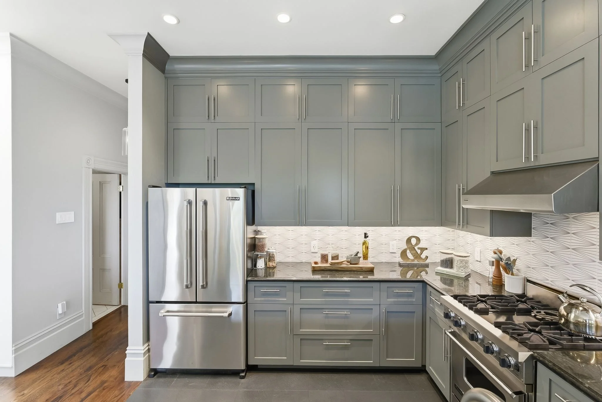 Modern kitchen with gray cabinets, stainless steel refrigerator, white patterned backsplash, stove, and kitchen utensils.