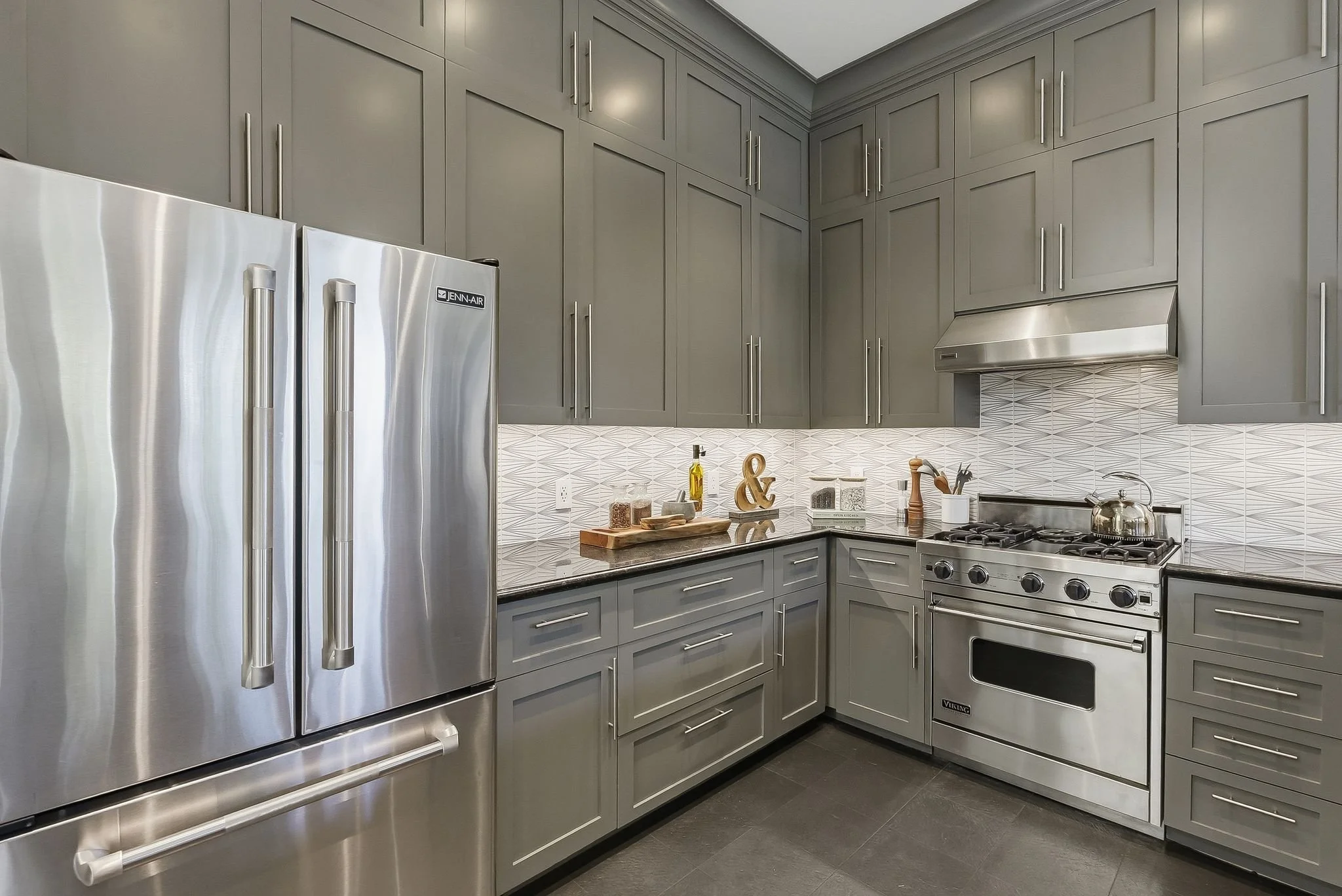Modern kitchen with gray cabinets, stainless steel refrigerator, oven, and range hood. Countertop with decorative items and kitchen tools.