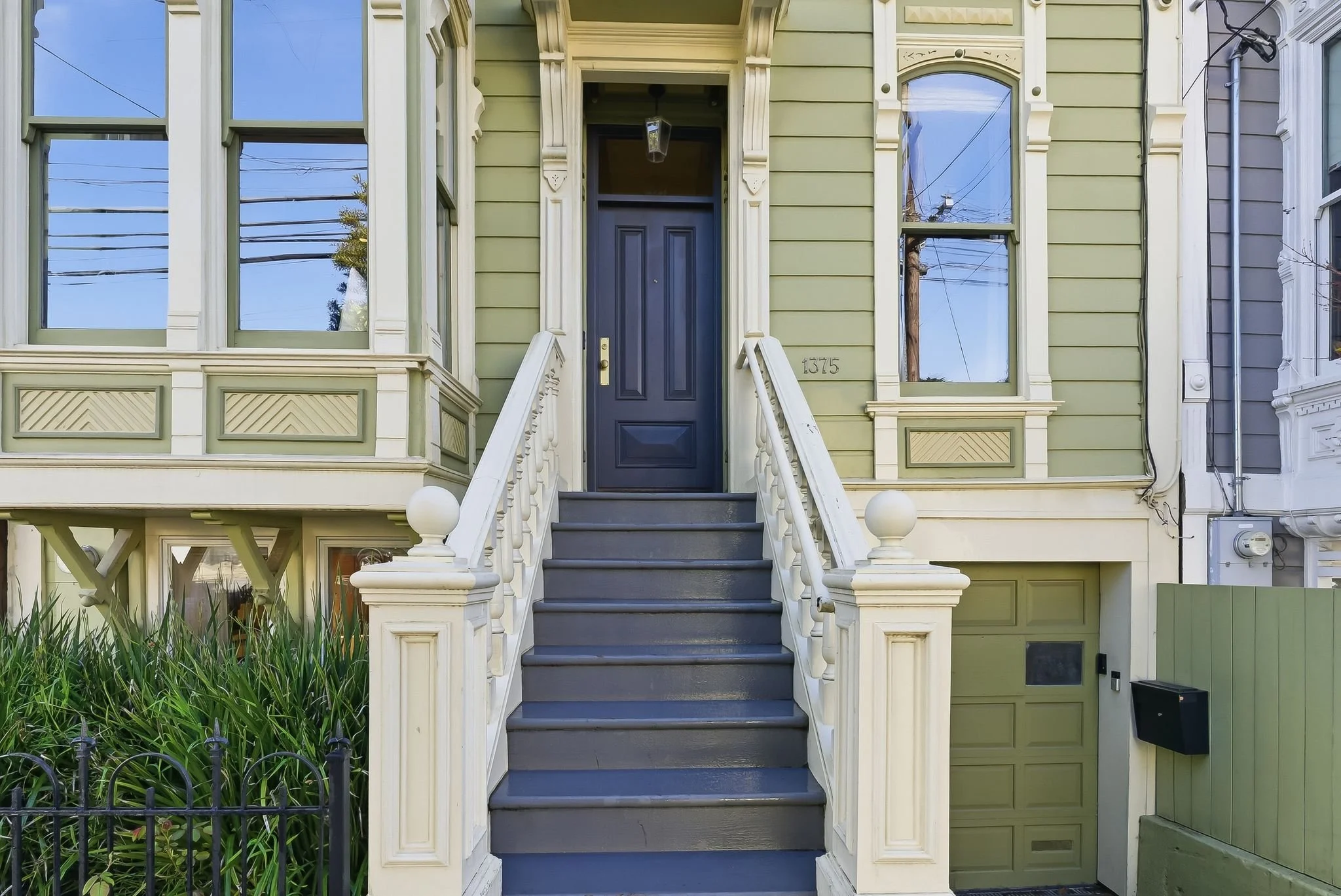 Front view of a two-story house with a staircase leading to a navy blue front door. The house has light green siding with ornate white trim and large windows. A small garden with green plants is on the left, bordered by a black fence. The house numbe
