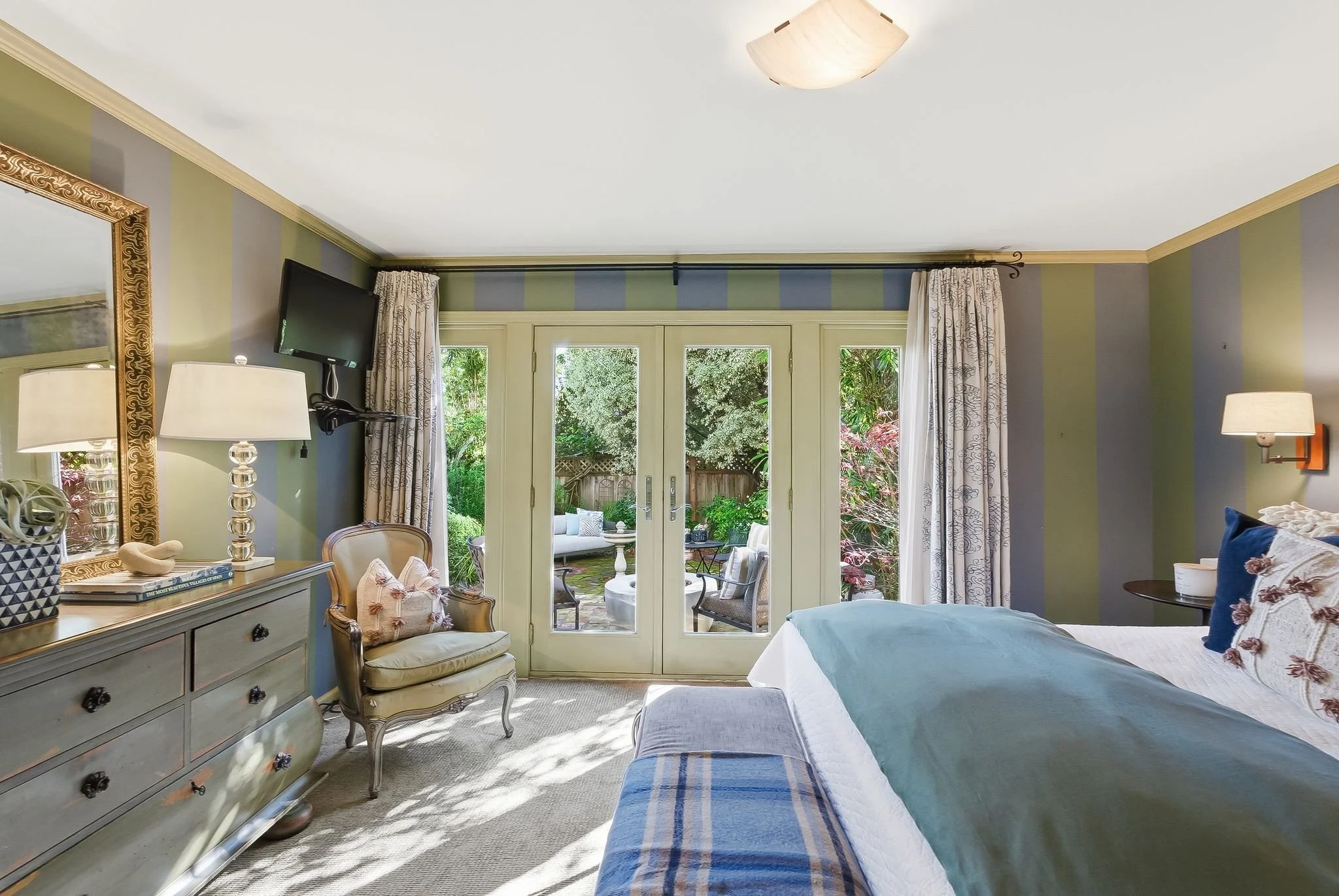A cozy bedroom with green and gray striped walls, a large bed with pillows and a blanket, a wooden dresser with a mirror, and a view of a lush garden outside through glass doors.