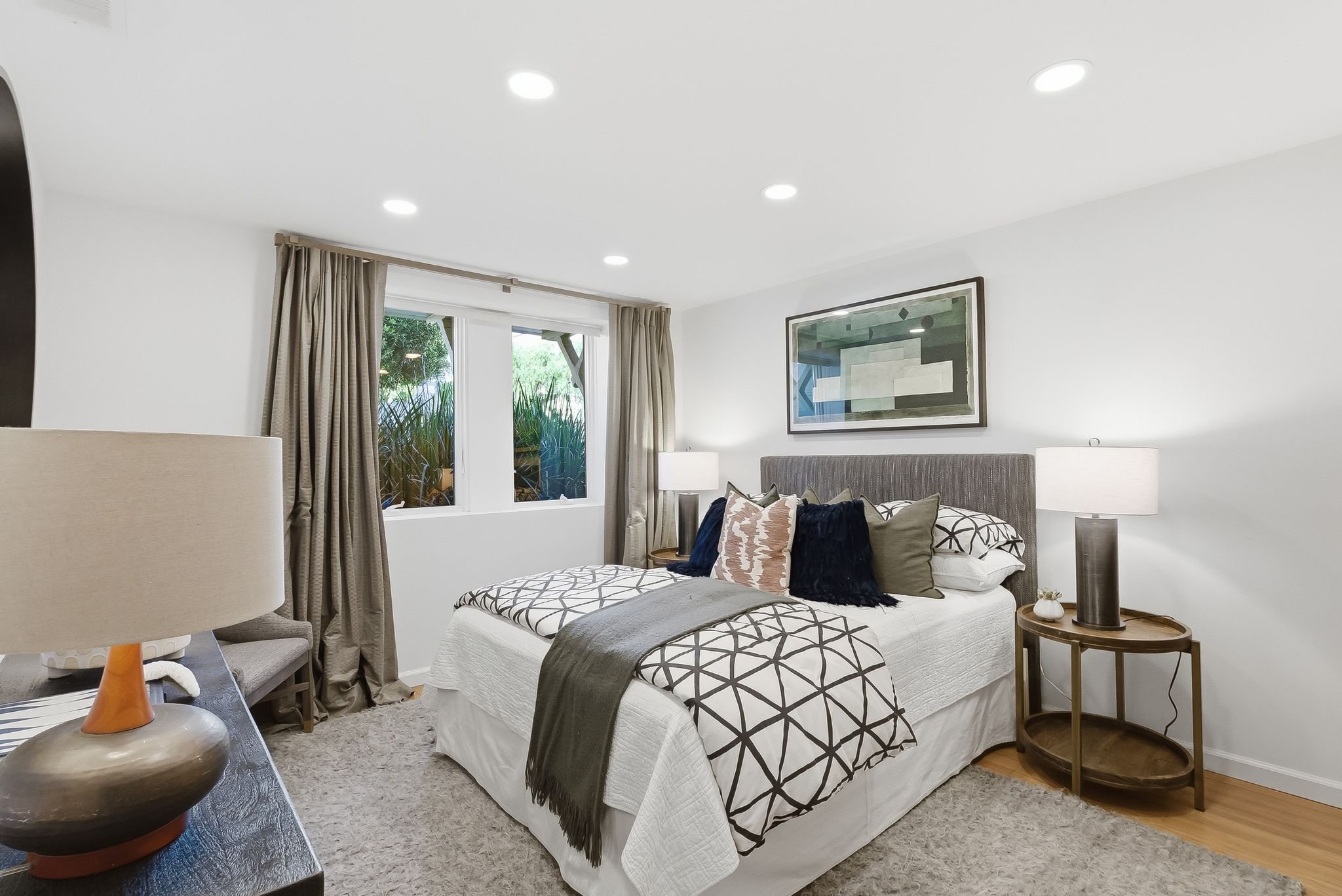 A bedroom with a bed, side table, lamps, window, and framed artwork.