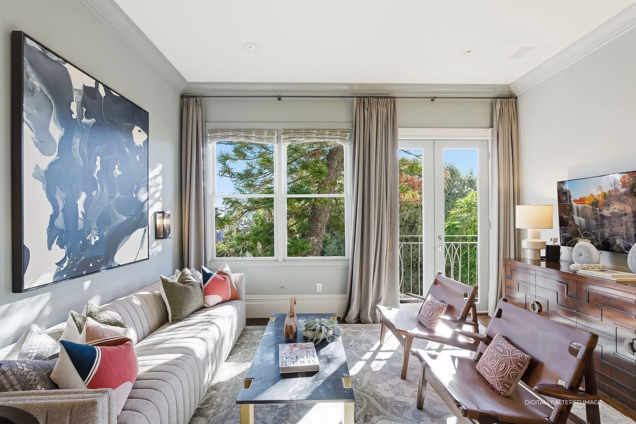 Living room with a beige sofa, colorful cushions, a black marble coffee table with decor, two brown leather chairs with patterned pillows, a wooden sideboard with a lamp and decorative objects, and large windows and a glass door leading to a balcony with trees outside.
