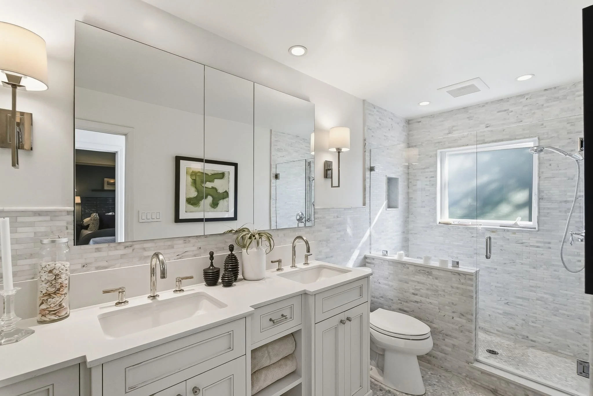 A modern bathroom featuring a double vanity with white cabinets, a large mirror, and decor items. The shower area has a glass door, a window, and white marble walls with a textured tile pattern.