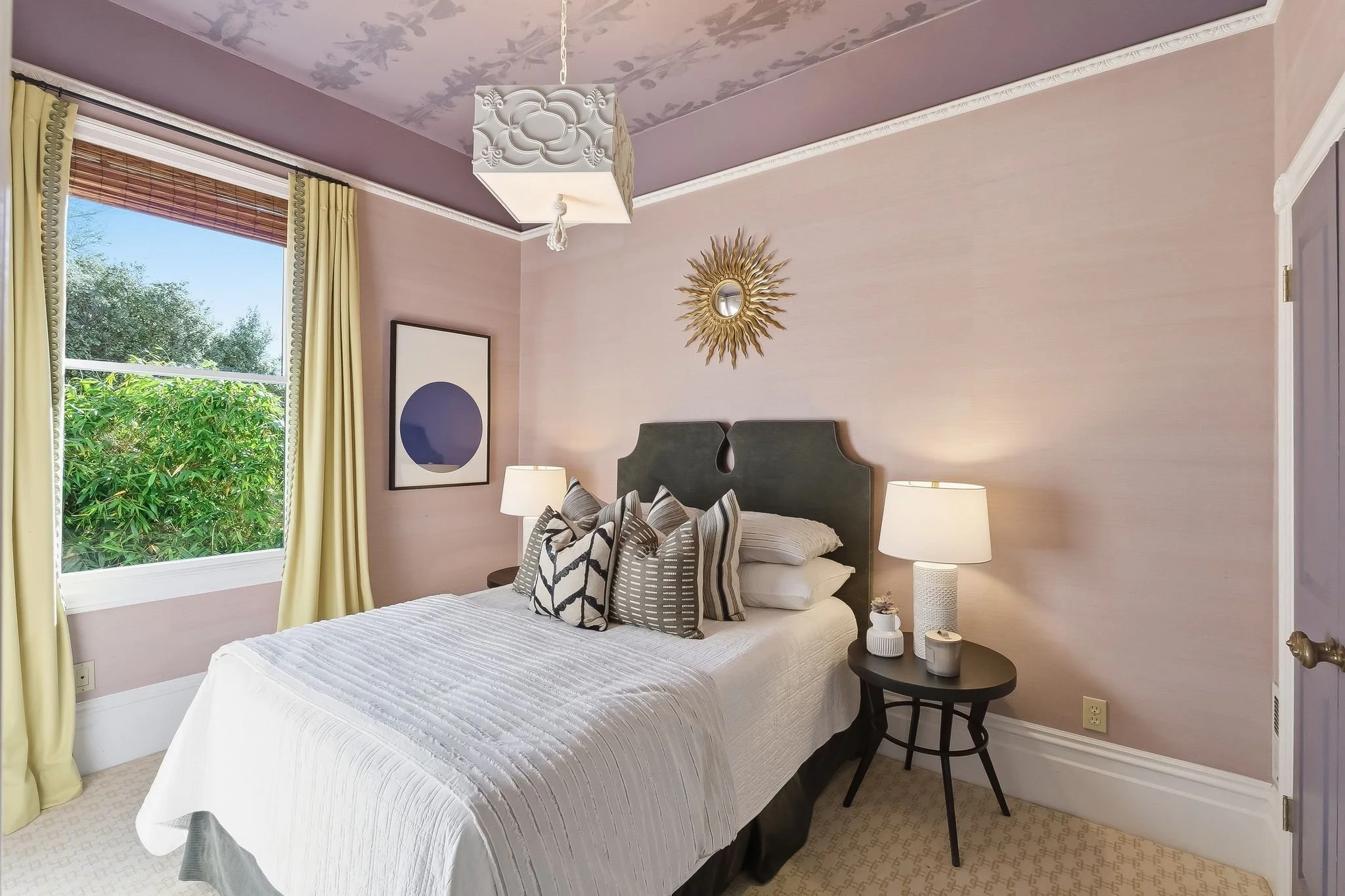 A cozy bedroom with a window showing greenery outside, a bed with decorative pillows, a black side table with a lamp and small decor items, a wall mirror with a sunburst frame, light pink walls, and cream curtains.