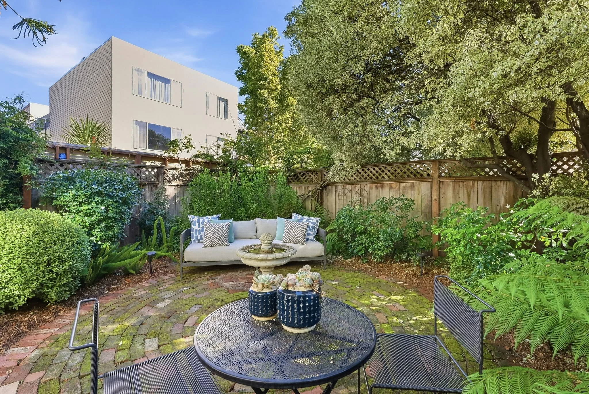 Backyard patio with brick paving, lush green plants, a white outdoor sofa with decorative pillows, black outdoor chairs, a round metal table with plants, and a wooden fence with a large tree.