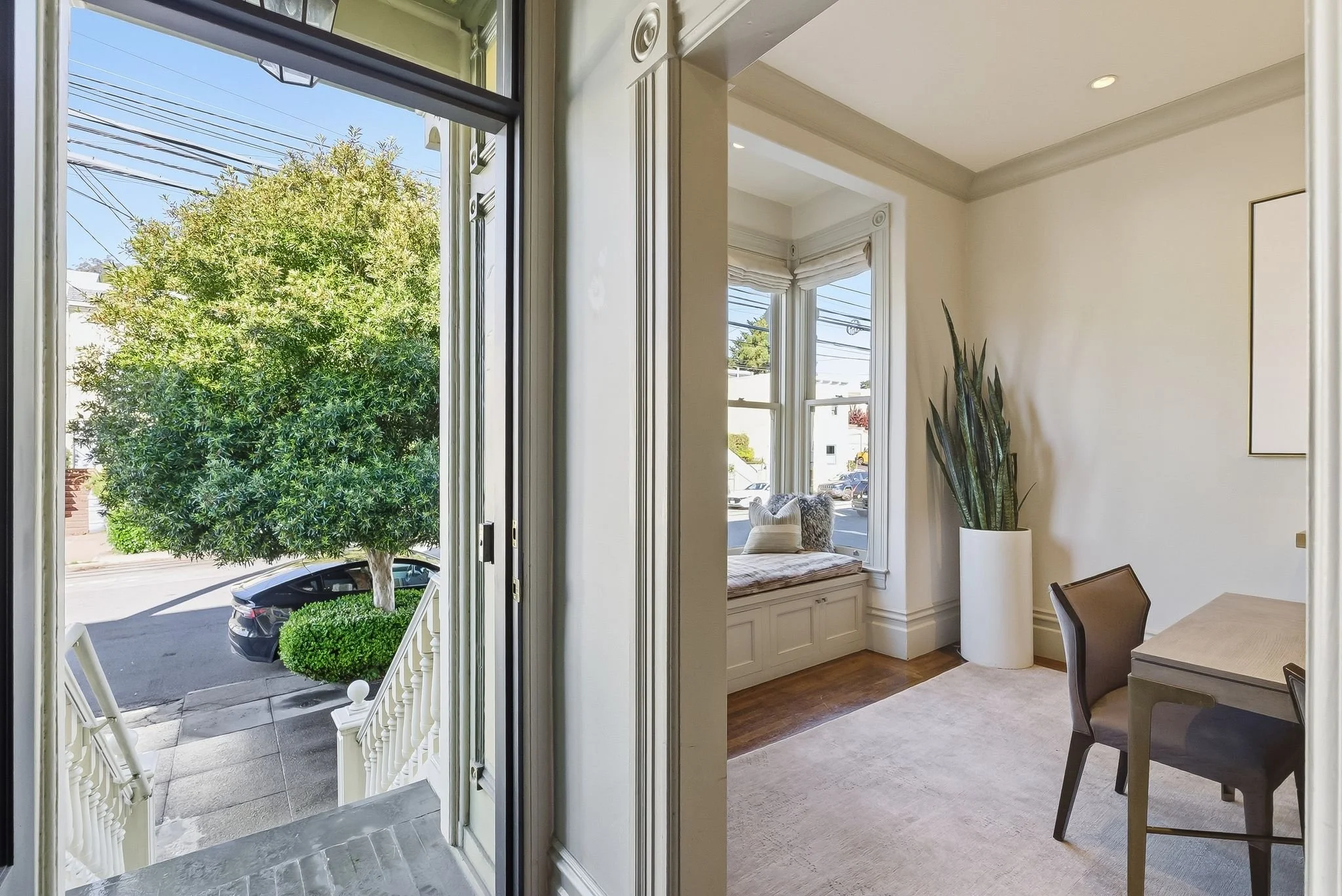 View from the front door of a home, looking through the doorway into a room with large windows, a padded window seat, a potted plant, and a desk with chairs, with sunny weather outside.