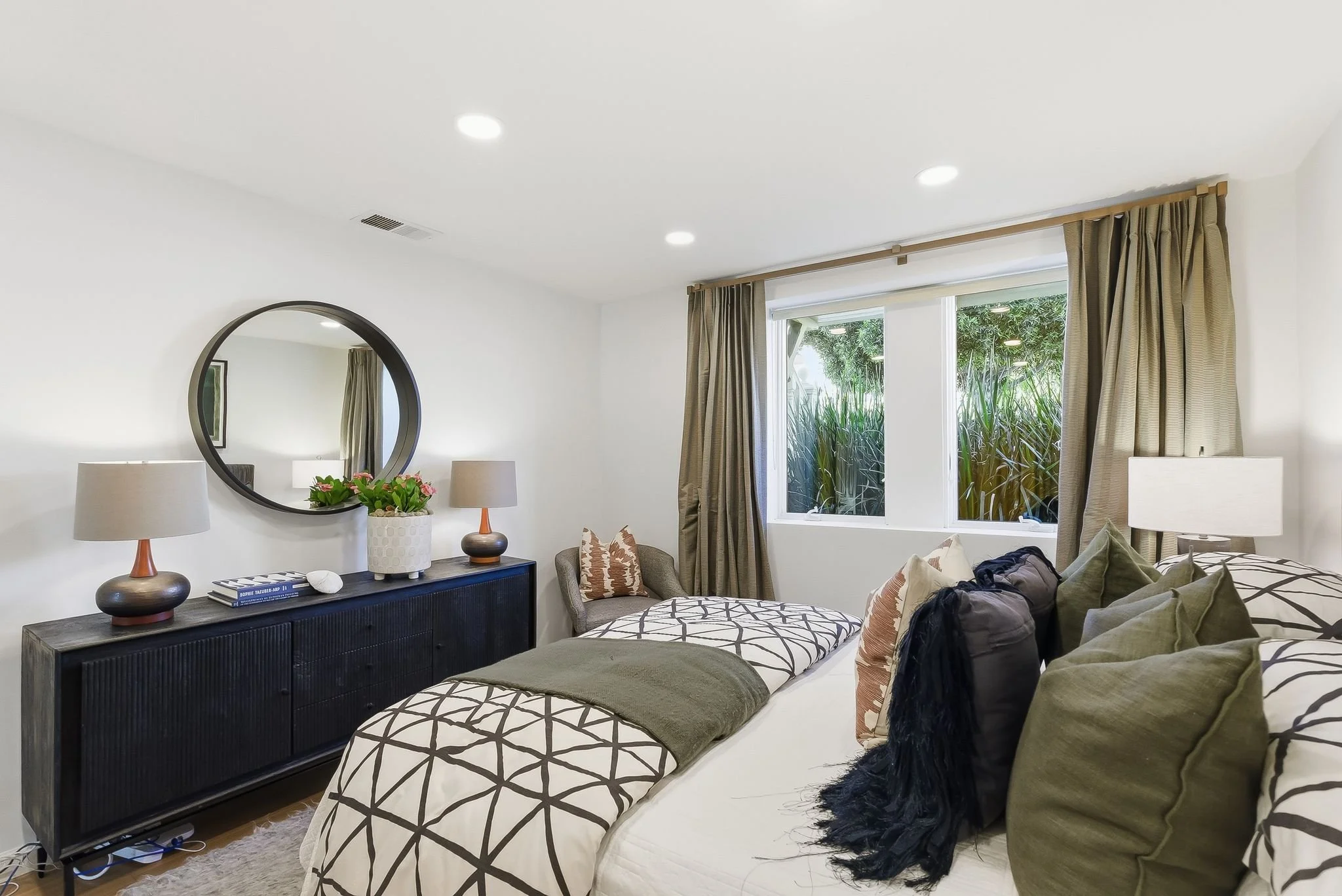 A bedroom with a bed piled with pillows, a side table with lamps, a dresser with a mirror, and large window with curtains overlooking greenery.