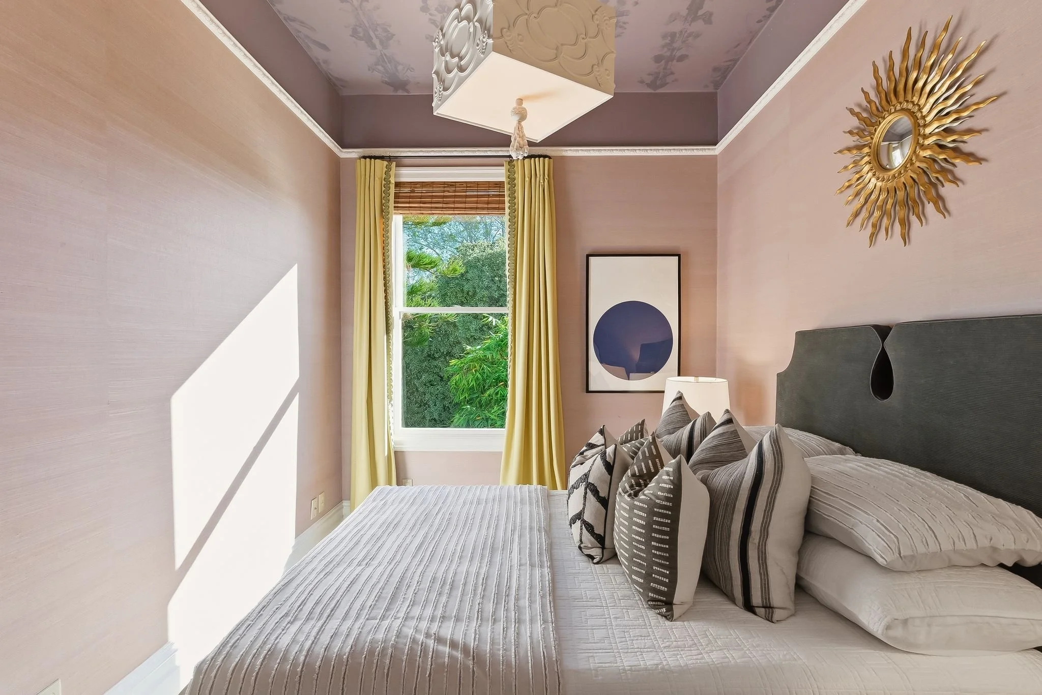 A cozy bedroom with a bed covered in white linens and multiple patterned pillows, a window with yellow curtains showing greenery outside, a framed artwork with a blue circle, a white lamp, and a gold sunburst mirror on the pink walls.