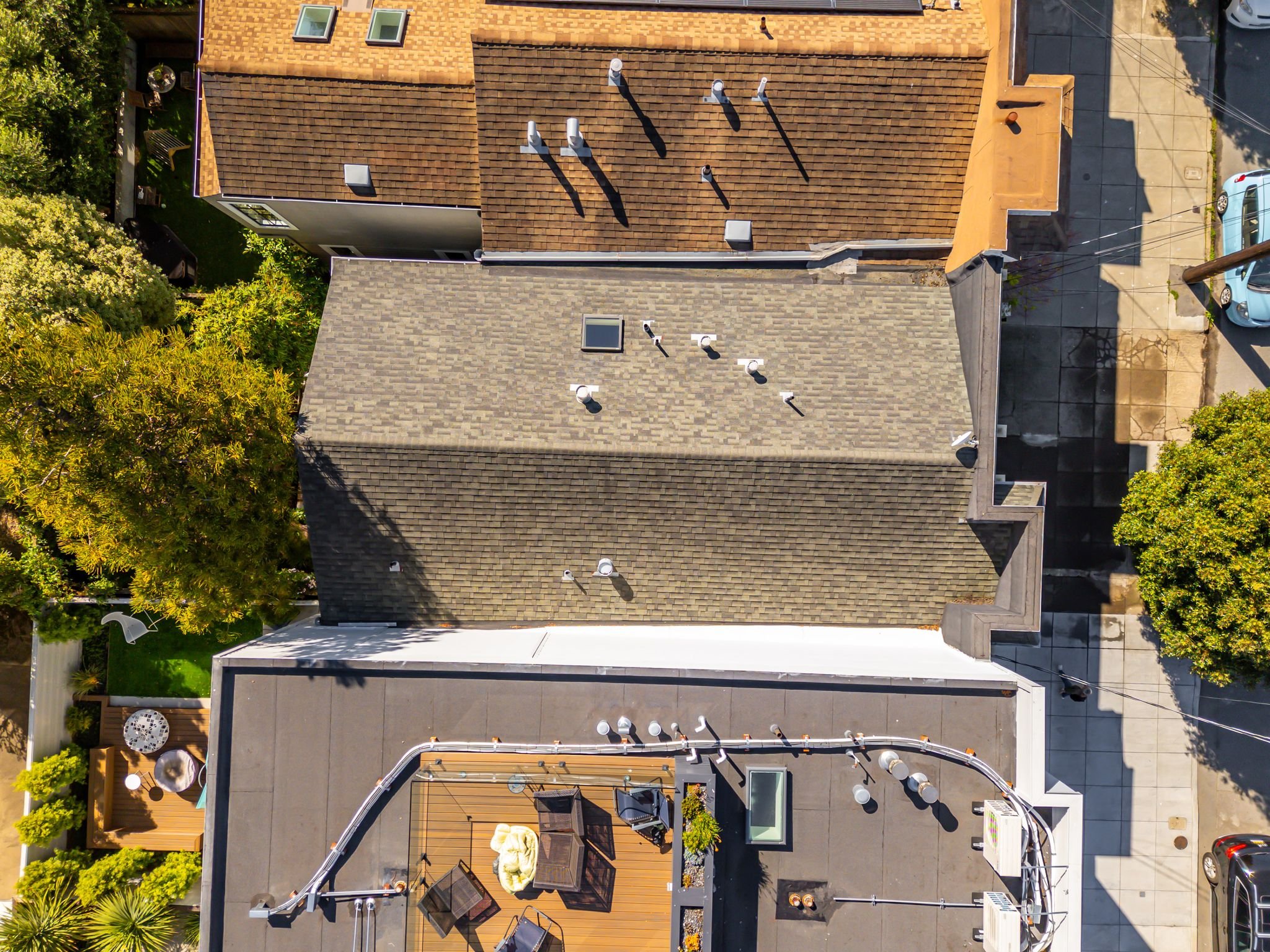 An aerial view of rooftops of several buildings with different colored shingles and flat rooftops, surrounded by trees, cars, and sidewalks.