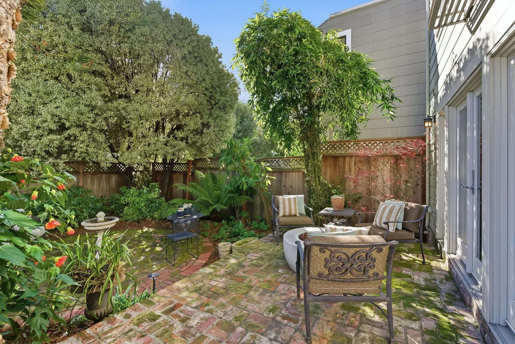 Cozy backyard patio with brick flooring, surrounded by lush green plants and trees, featuring outdoor seating with striped and patterned cushions, decorative pillows, and garden accessories.