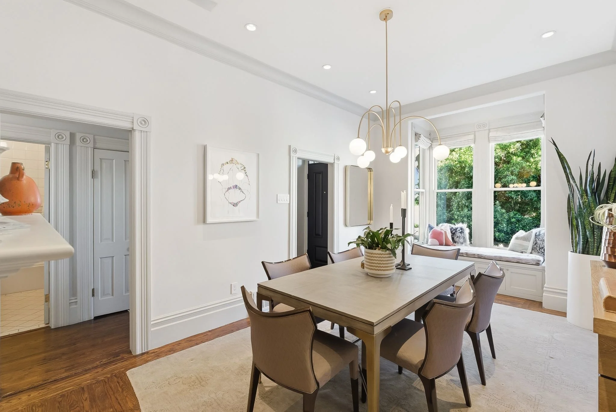 Bright dining room with a beige wooden table, six beige cushioned chairs, a modern chandelier, a window seat with cushions, and large windows with greenery outside.
