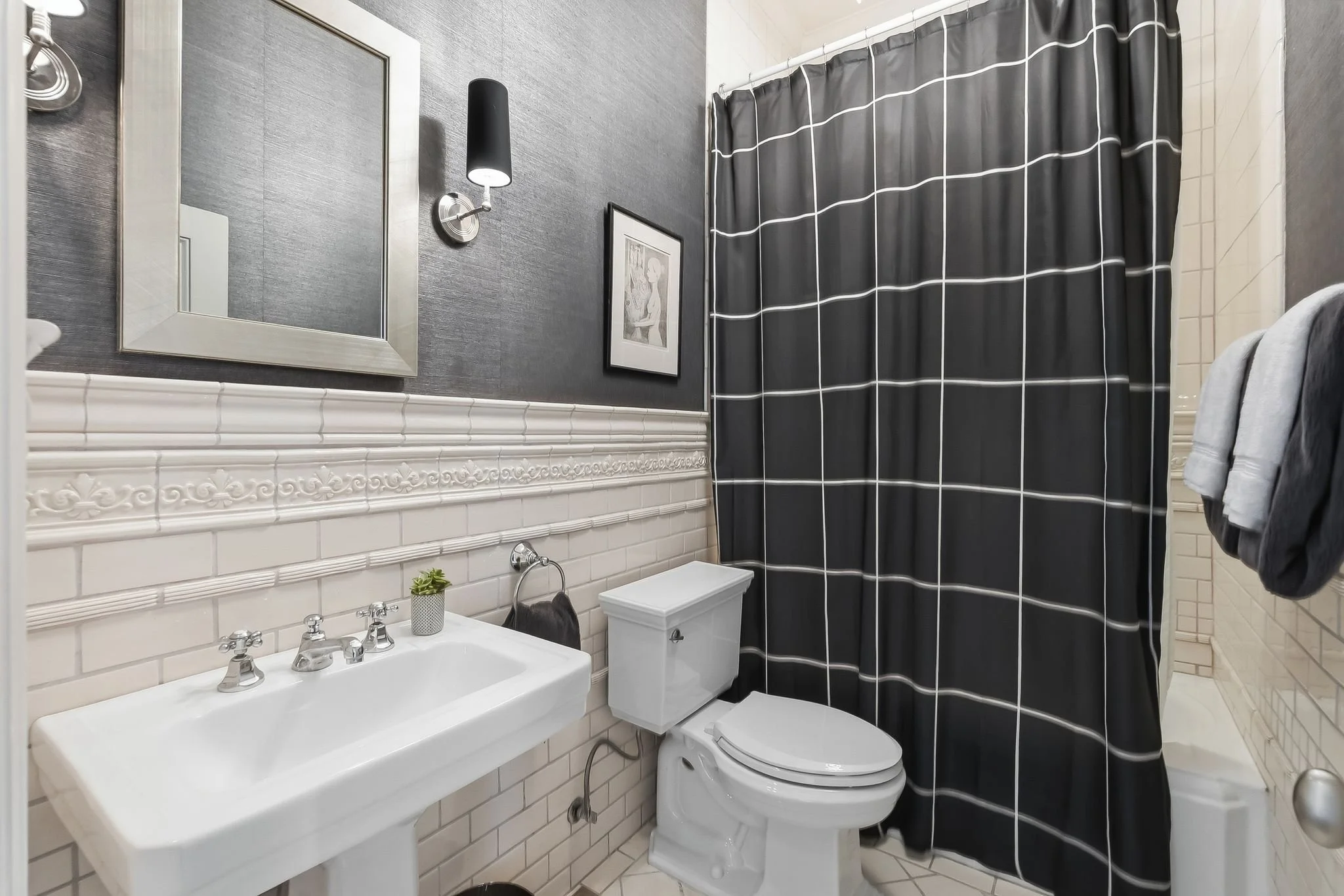 Small bathroom with black and white decor, featuring a pedestal sink, a toilet, a large mirror, dark wall with black wallpaper, a framed picture, a black and white checkered shower curtain, and a towel rack.