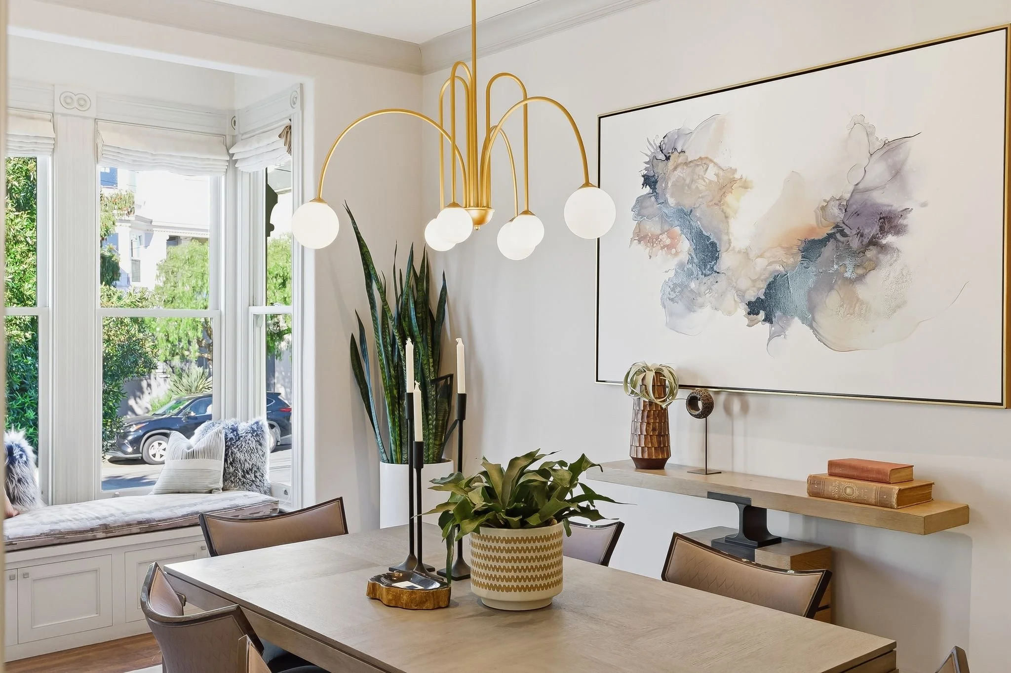Bright dining room with a window seat, round dining table, modern chandelier, large abstract wall art, green potted plant, and decorative vases.