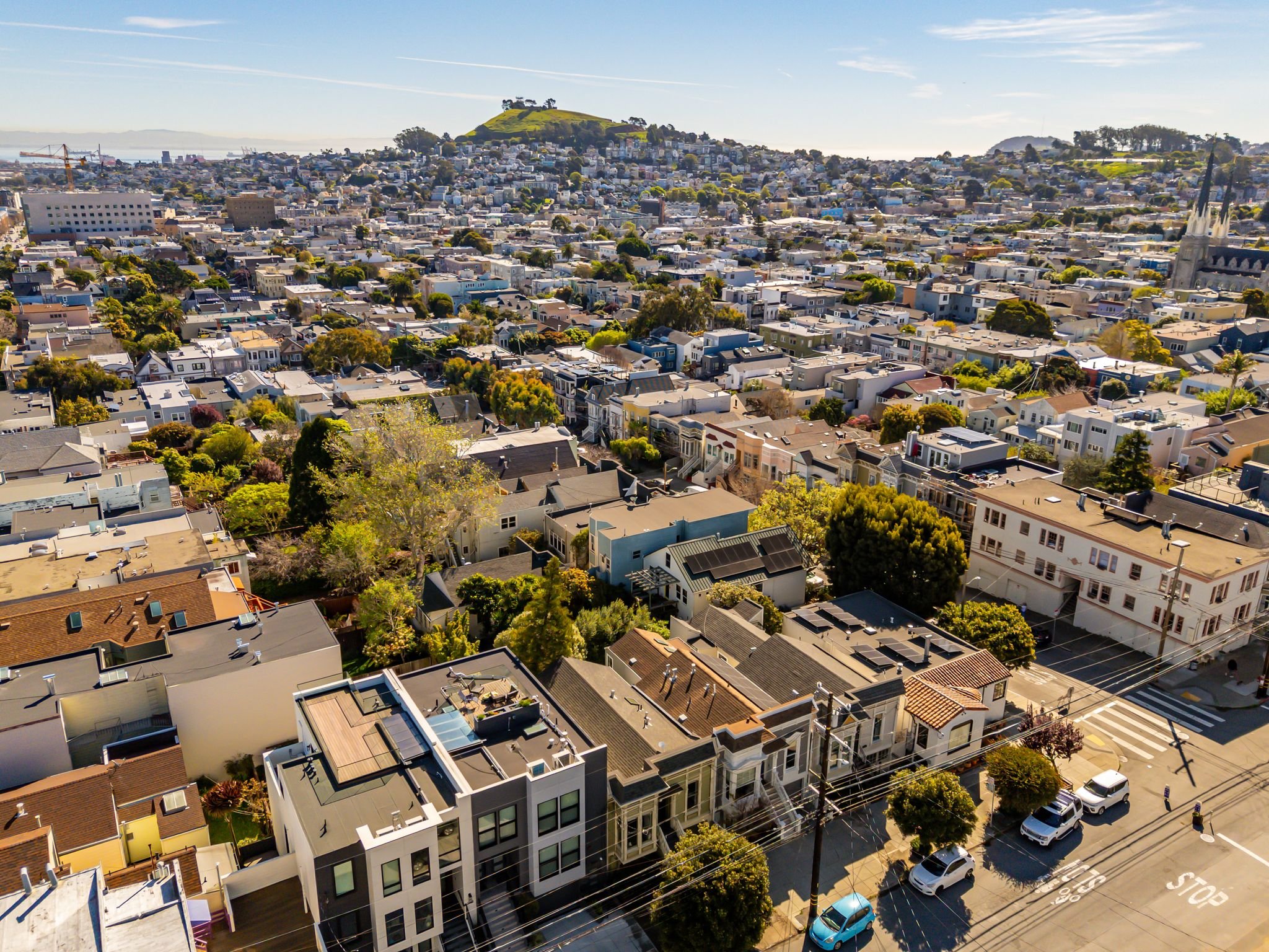 Aerial view of a city with numerous houses, trees, and streets, with a green hill in the background under a clear blue sky.