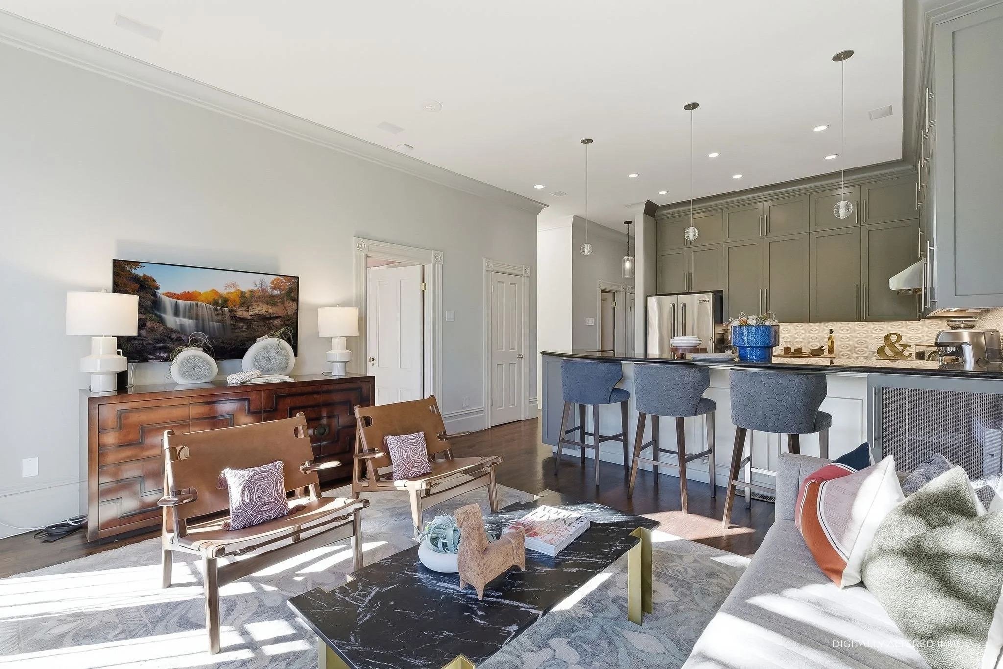 Living room with a black marble coffee table, beige sofa with pillows, wooden chairs with pillows, a wooden sideboard with lamps and decor, and a kitchen with gray cabinets, a breakfast bar with four chairs, stainless steel appliances, and pendant lighting.