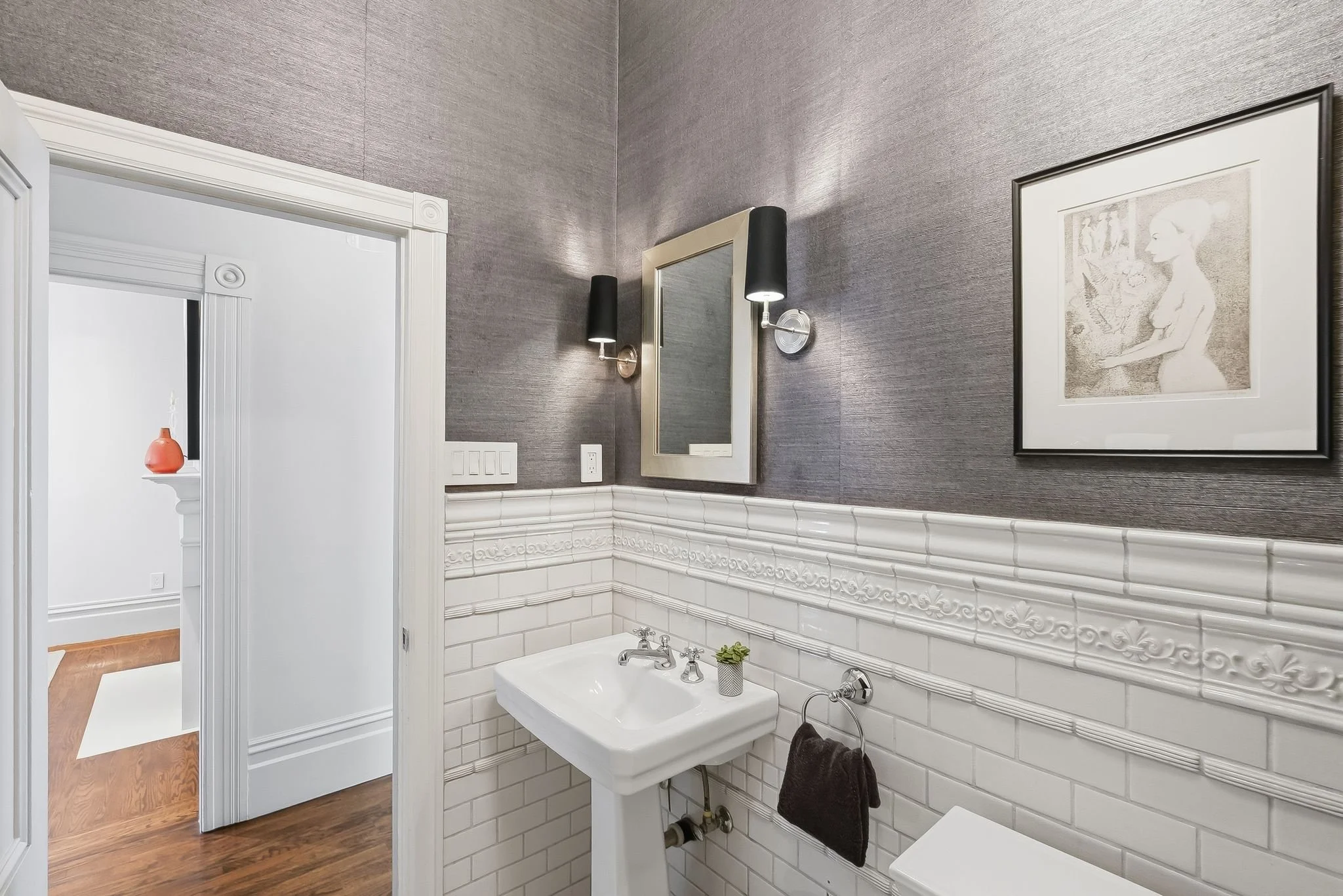 Bathroom with dark textured wall, white ceramic tile wainscoting, a rectangular mirror, modern black wall sconces, framed artwork of a woman, white pedestal sink, small plant, black towel on a ring, and nearby doorway revealing part of another room w