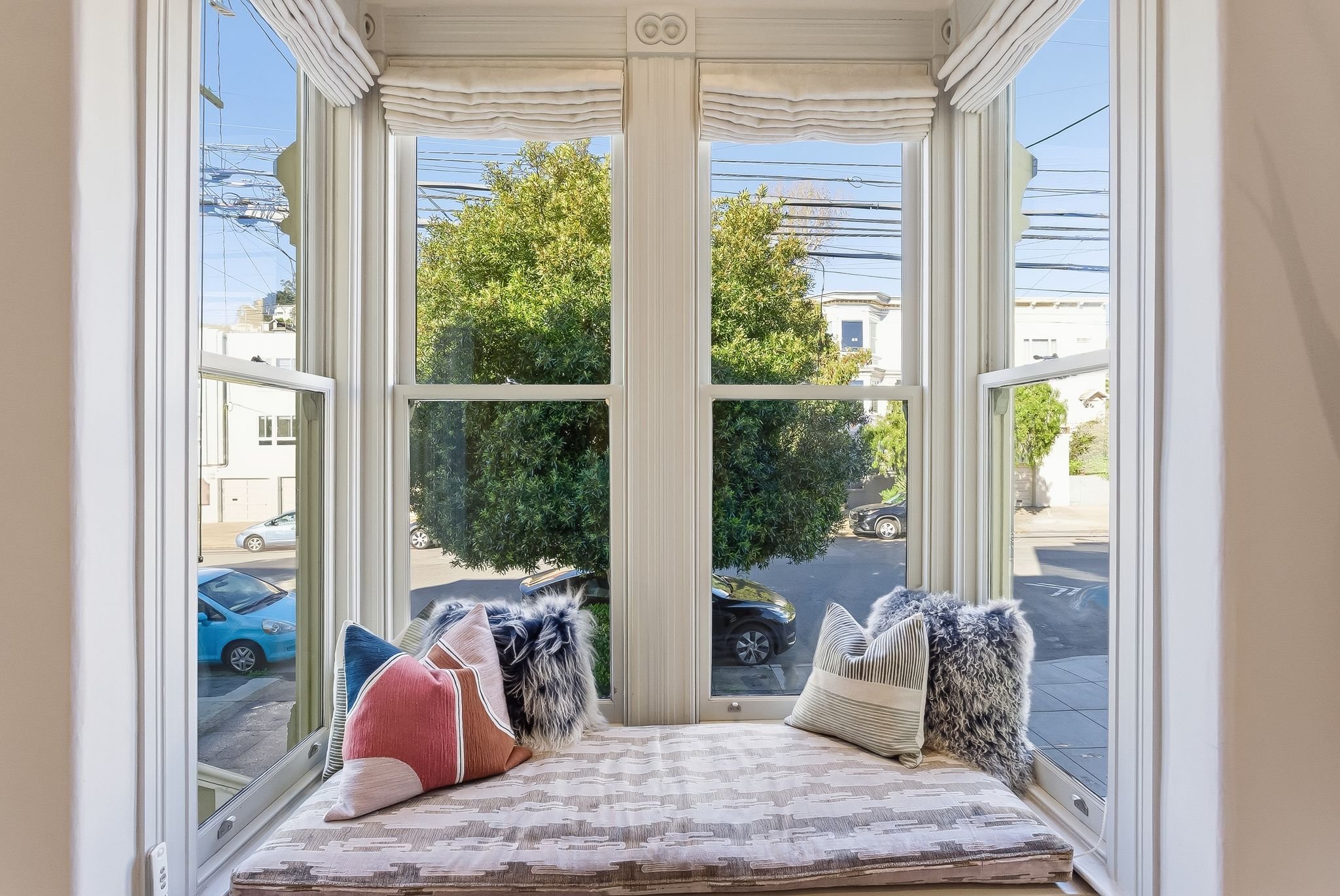 A window nook with a cushioned seat and decorative pillows overlooking a street with parked cars and trees, with power lines visible above.