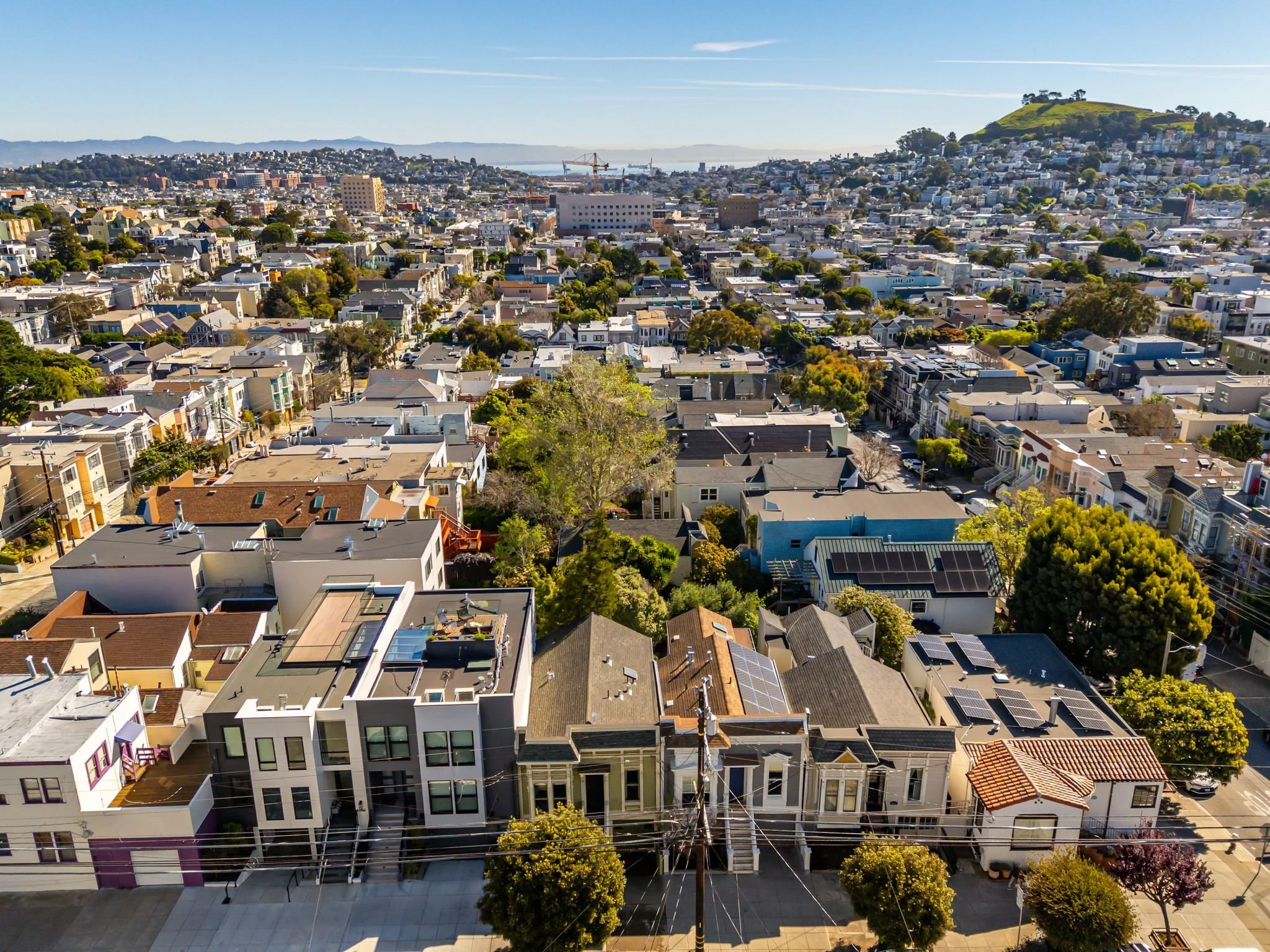 Aerial view of a San Francisco neighborhood with a mix of modern and historic homes, many with solar panels on roofs, trees lining streets, and hills in the background.